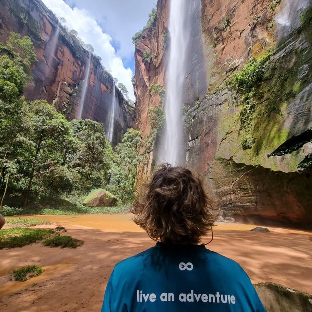Standing under five waterfalls at once is a good problem to have. A traveler in a teal 'live an adventure' shirt gazes up at multiple waterfalls cascading down tall red sandstone cliffs into an ochre-colored pool during a Jalapão adventure hike