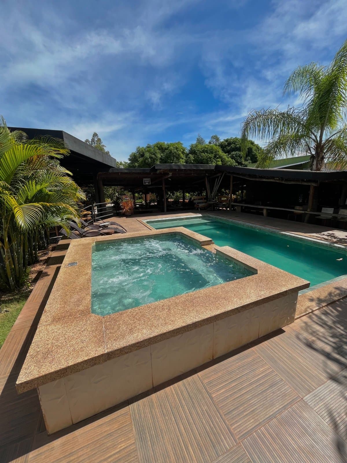 After a full day on the trails, this is exactly where you want to be. Outdoor jacuzzi and swimming pool with turquoise water at a Jalapão adventure lodge, surrounded by palm trees and a shaded pergola under a blue sky