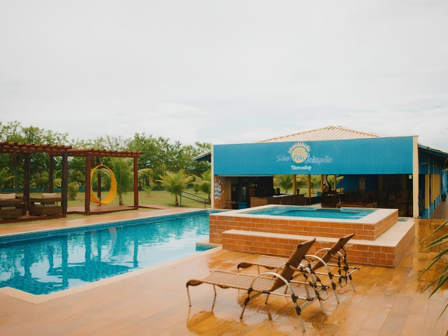 Cold beer, warm water, and nobody fighting over the sun loungers yet. Outdoor pool and elevated hot tub at Pousada São Félix do Tocantins, with wicker sun loungers on a tiled deck, a yellow hanging chair under a pergola, and the blue-fronted guesthouse building in the background — a comfortable base for a Jalapão adventure