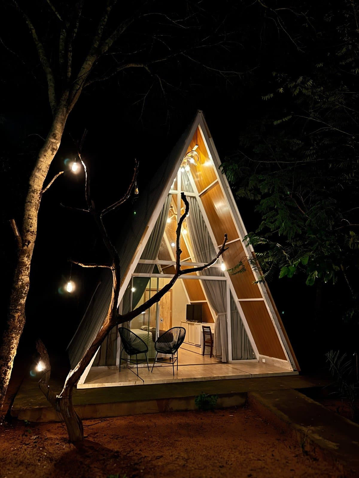 After a day on the trail, this is exactly the welcome you want. An illuminated A-frame cabin glowing with warm light at night, surrounded by dark forest trees and string lights, with two chairs on an open timber deck in a Jalapão trekking lodge