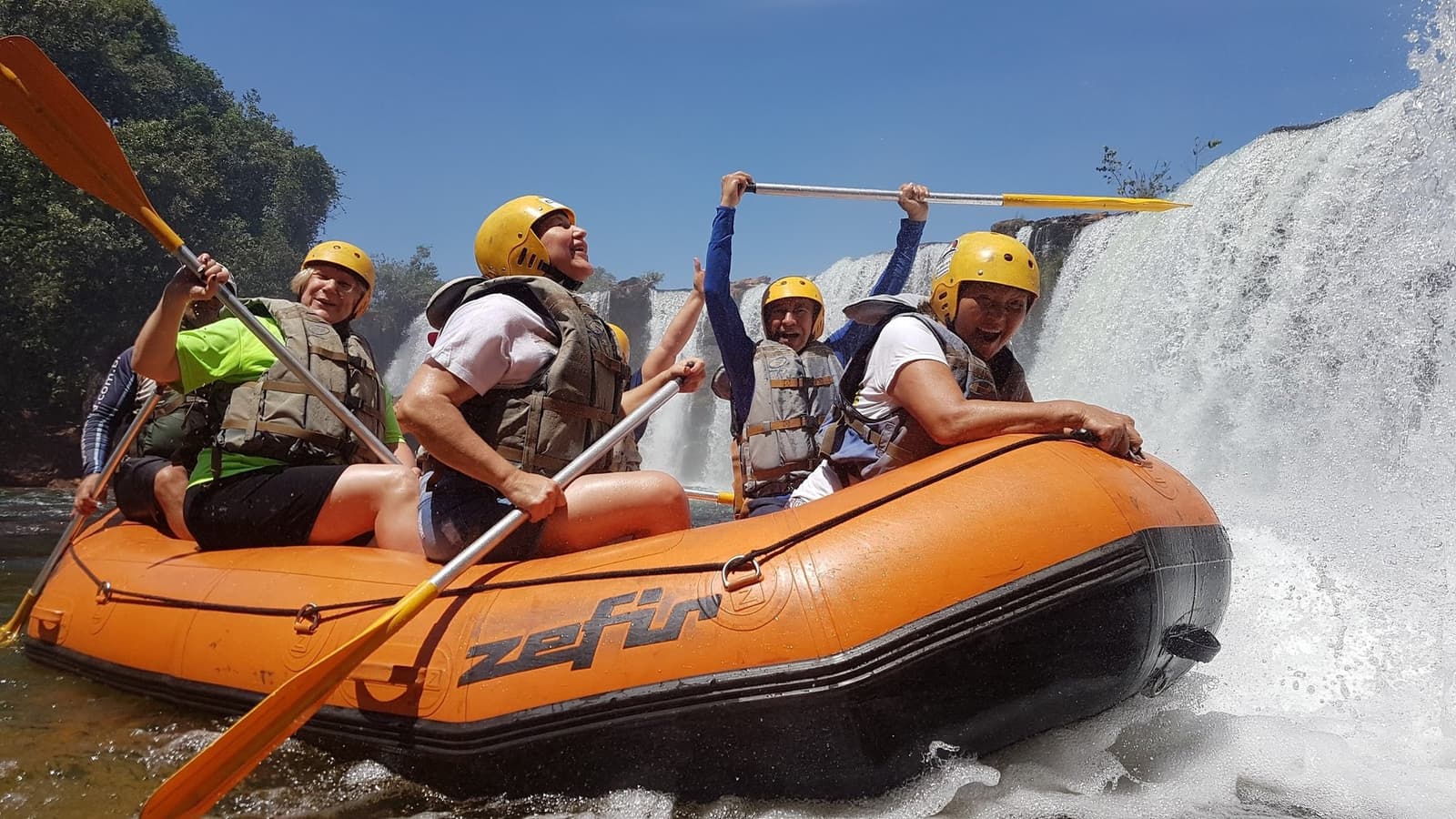 Paddles up, faces soaked, the Novo River has opinions about who's in charge. Five smiling rafters in yellow helmets and life jackets paddle an orange inflatable raft past a powerful waterfall on a Jalapão trekking and rafting adventure