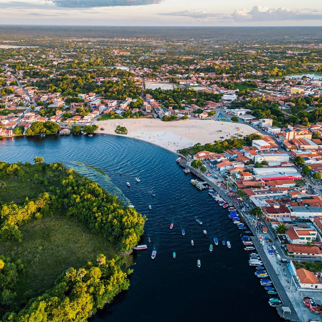 The town wraps around the river like it has nowhere better to be. Aerial view of Barreirinhas showing the Preguiças River curving past a white sand beach, colorful boats moored along the waterfront, and the town spreading into green vegetation on the Lençóis Maranhenses Full Itinerary