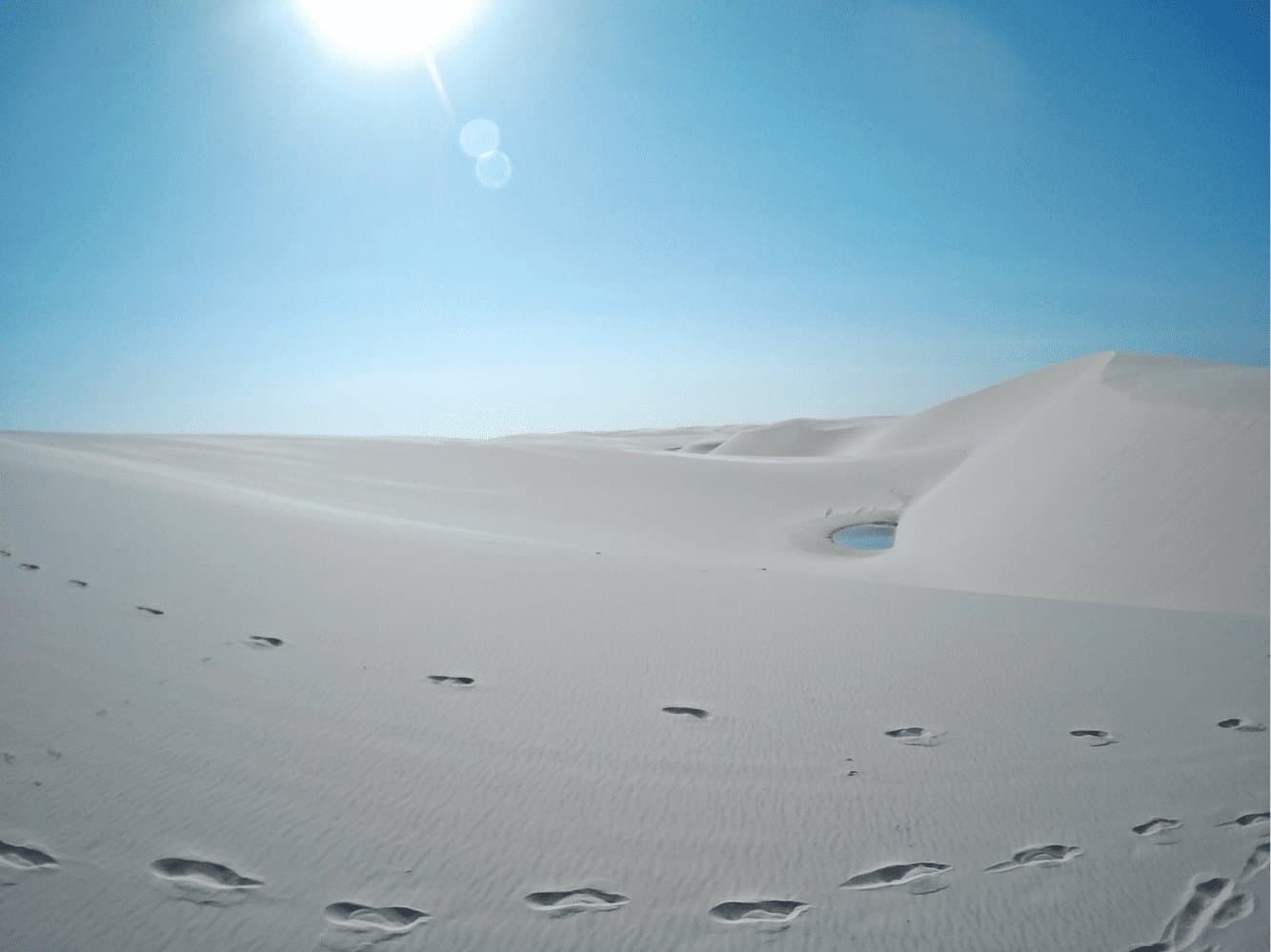 Someone walked this way, found water, and probably couldn't believe their luck. Bare footprints crossing vast white sand dunes toward a small blue lagoon nestled between ridges under a blazing sun — a scene from the Lençóis Maranhenses Full Itinerary