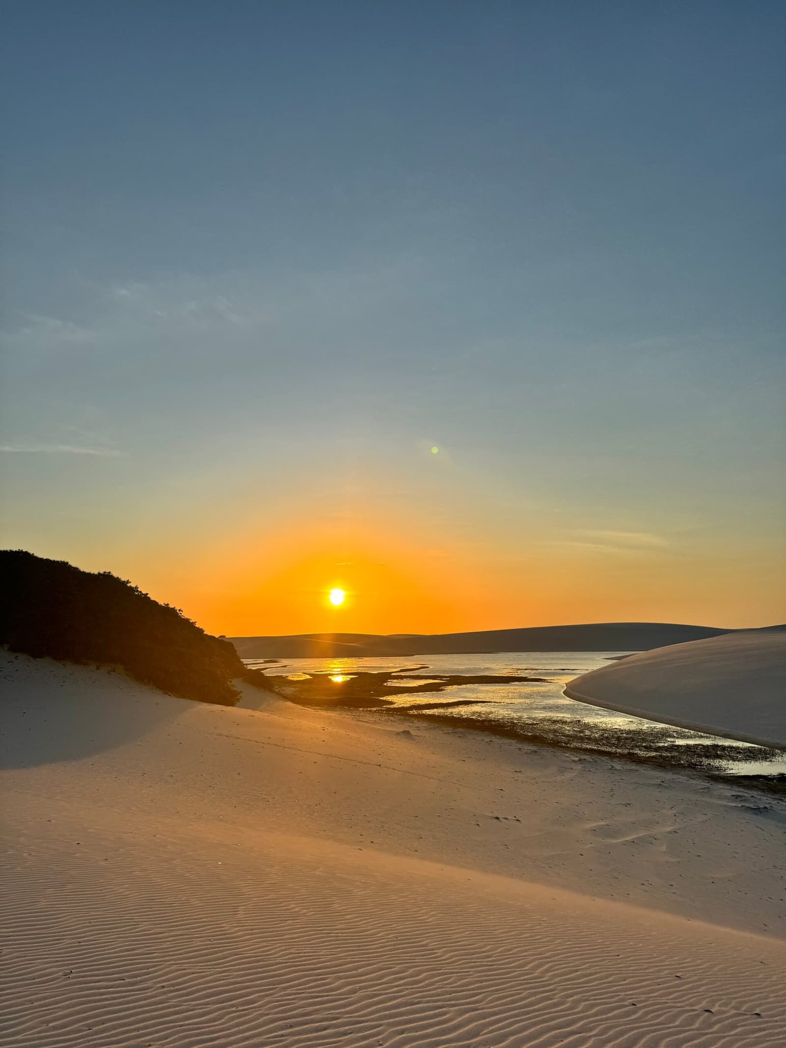 The dunes hold the last light like they're not ready to let go. Golden sunset over a tidal channel winding between white sand dunes in Lençóis Maranhenses, reflecting orange light on shallow water
