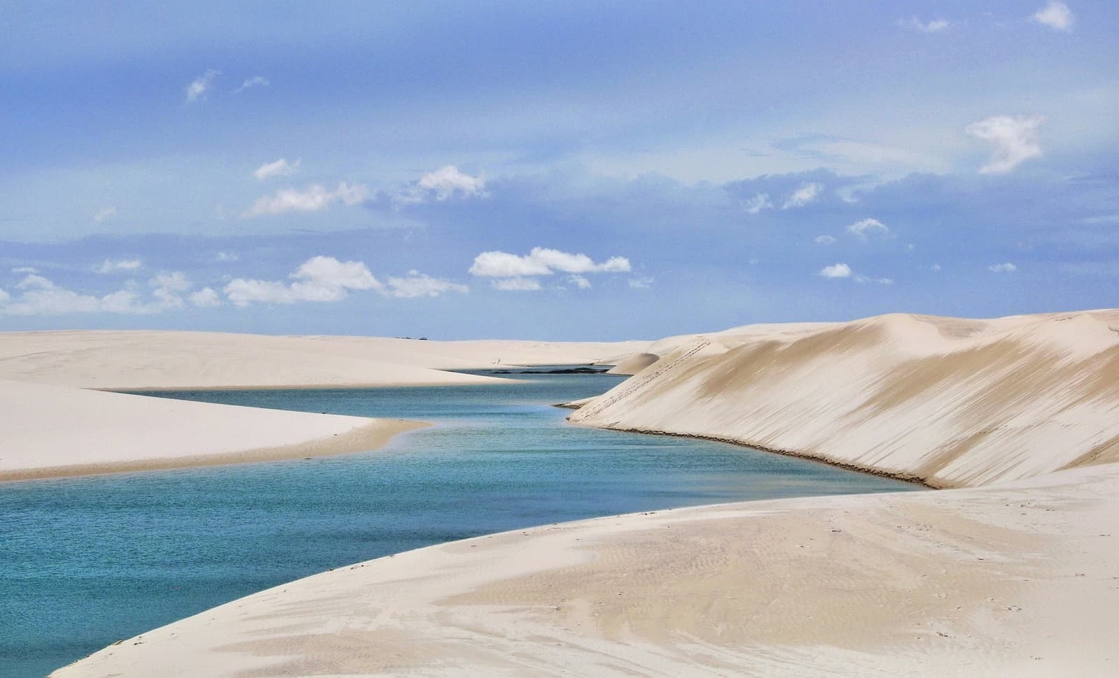 Fresh water, white sand, blue sky, and no two days look quite the same. Turquoise rainwater lagoon nestled between sweeping white sand dunes under a blue sky in Lençóis Maranhenses National Park