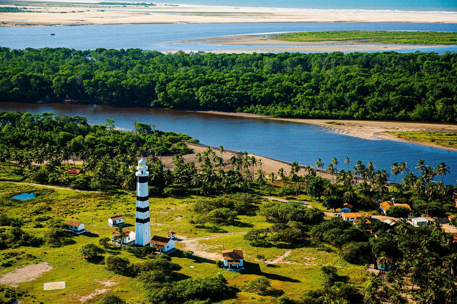 The lighthouse that has watched rivers shift and sandbanks come and go for generations. Aerial view of the black-and-white striped Mandacaru lighthouse surrounded by palm trees and dense forest, with a wide river, sandy banks, and white dunes on the horizon — the starting landscape of the Lençóis Maranhenses hike
