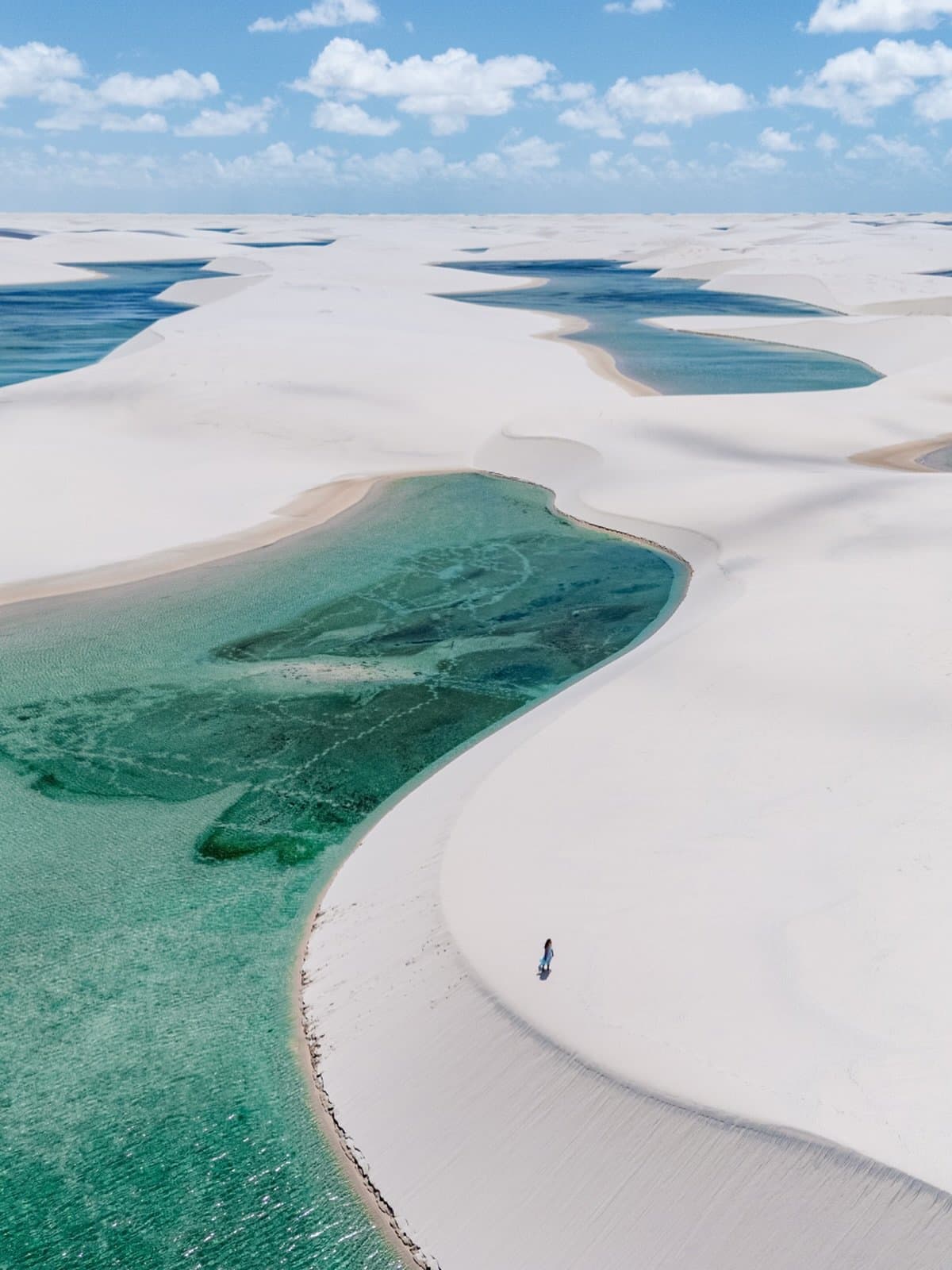 Just you, the dunes, and a lagoon that looks painted on. Aerial view of Lençóis Maranhenses Santo Amaro, showing sweeping white sand dunes dotted with turquoise and emerald rainwater lagoons stretching to the horizon, with a single person standing at the water's edge under a blue sky