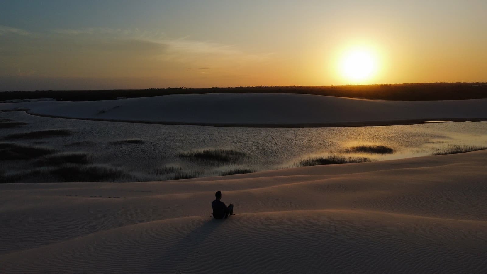 Some sunsets you share. This one you keep. A person sitting alone on a white sand dune in Lençóis Maranhenses Santo Amaro, watching the sun set over a still lagoon fringed with grasses and distant tree line