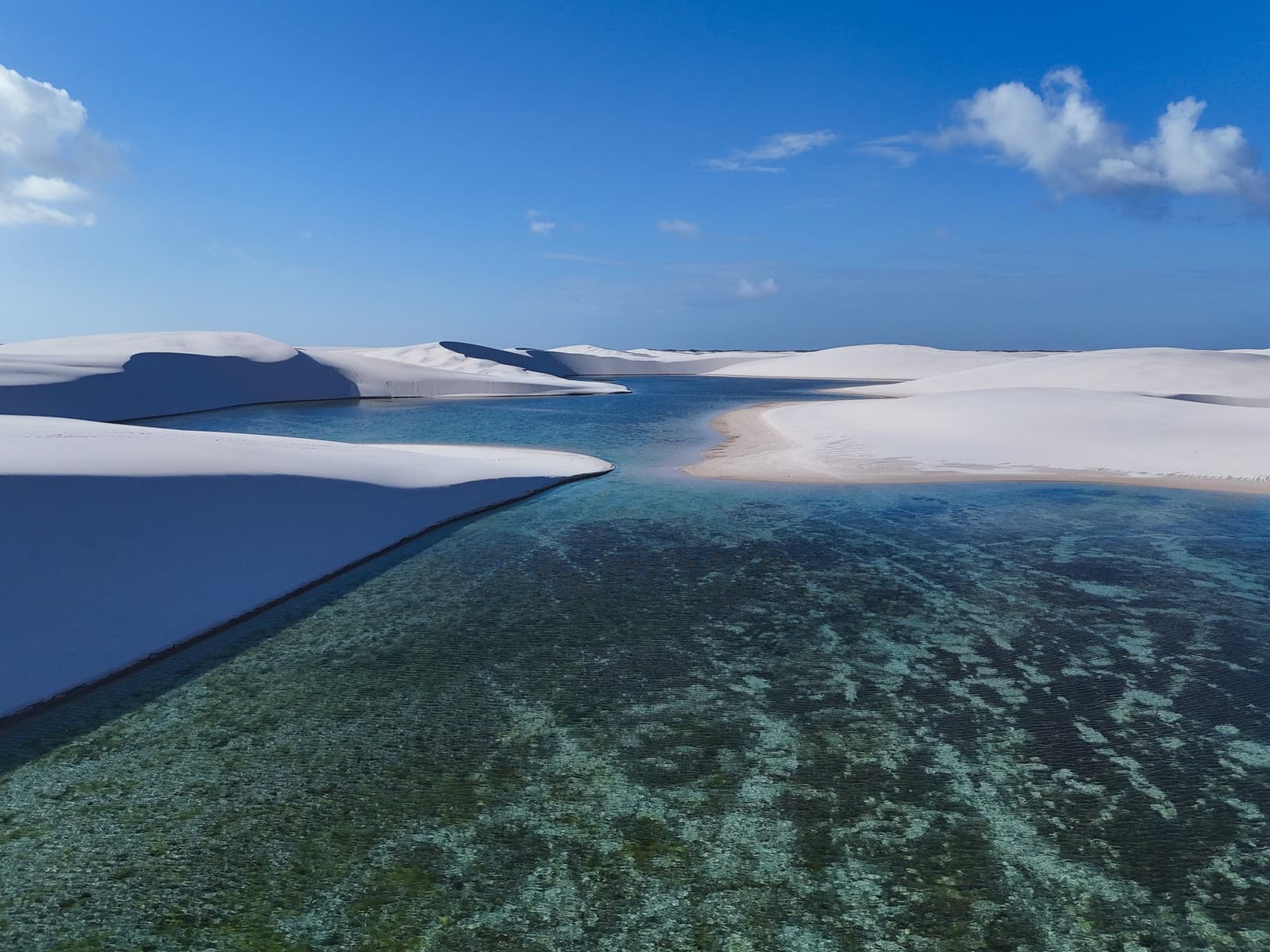 Fresh water, white sand, blue sky, Brazil keeps a few secrets worth trekking for. Aerial view of a clear green-tinted freshwater lagoon cutting between sculpted white sand dunes under a blue sky in Lençóis Maranhenses National Park, with the water so transparent the sandy bottom is fully visible