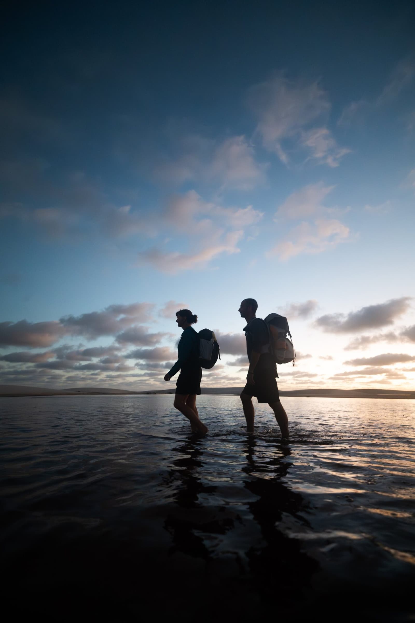 Wet feet, full packs, and a sky that earns it. Two hikers with backpacks silhouetted against a dusk sky, wading barefoot through shallow water on the lencois maranhenses trek