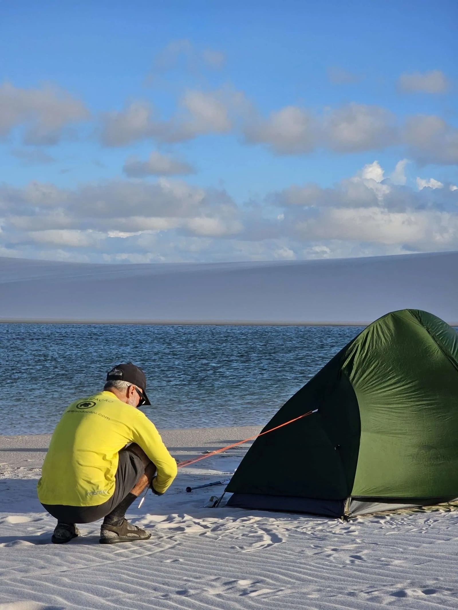 Home for the night sorted, the dunes can wait till morning. A guide in a yellow shirt crouches to stake down a green dome tent on white sand beside a calm blue lagoon during a Lençóis Maranhenses trekking expedition, with vast dunes visible across the water