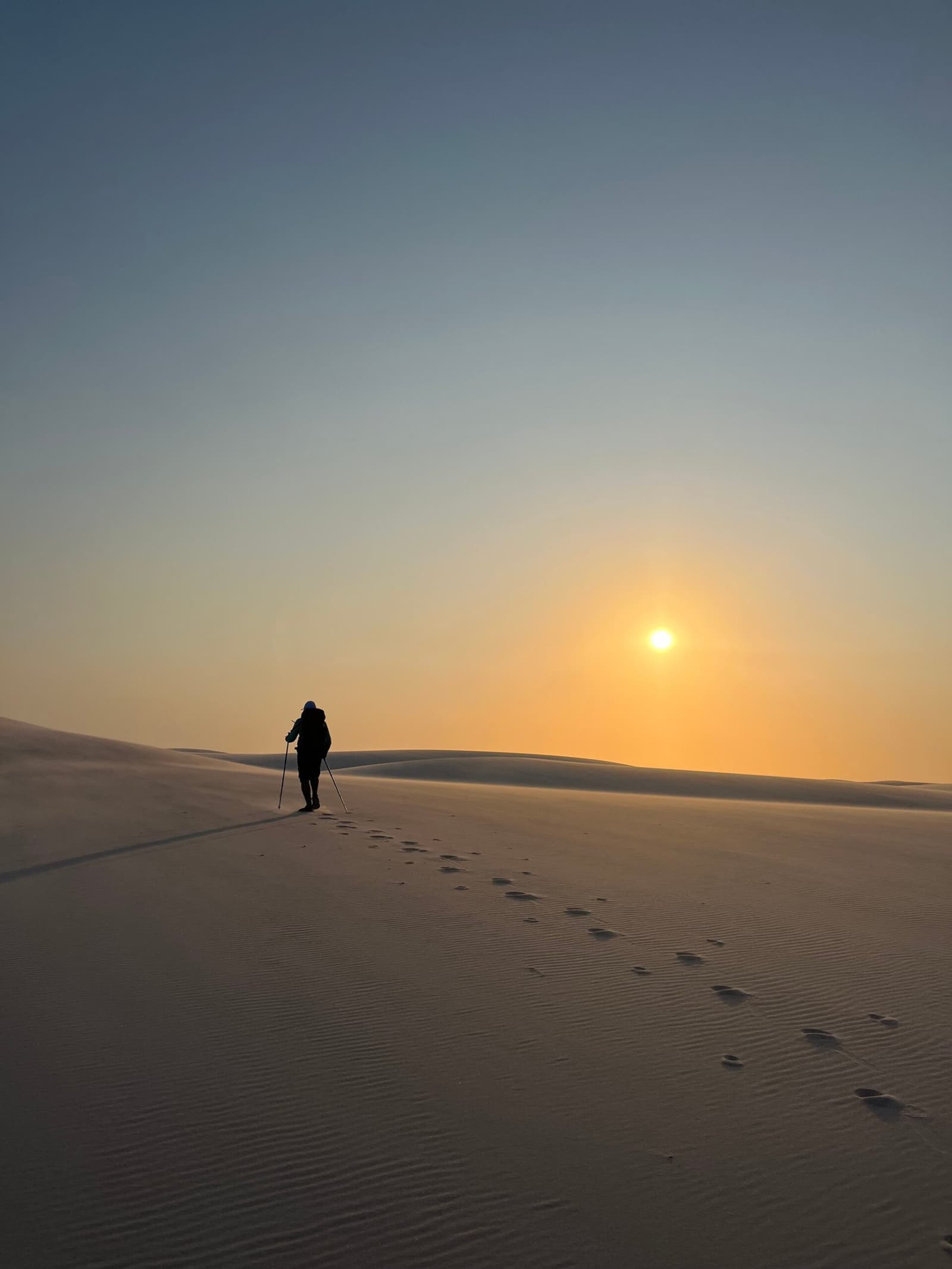 One set of footprints, one horizon, zero wrong turns. A lone trekker with hiking poles climbing a vast white sand dune during Lençóis Maranhenses trekking, leaving a trail of footprints behind as the sun rises low on the golden horizon