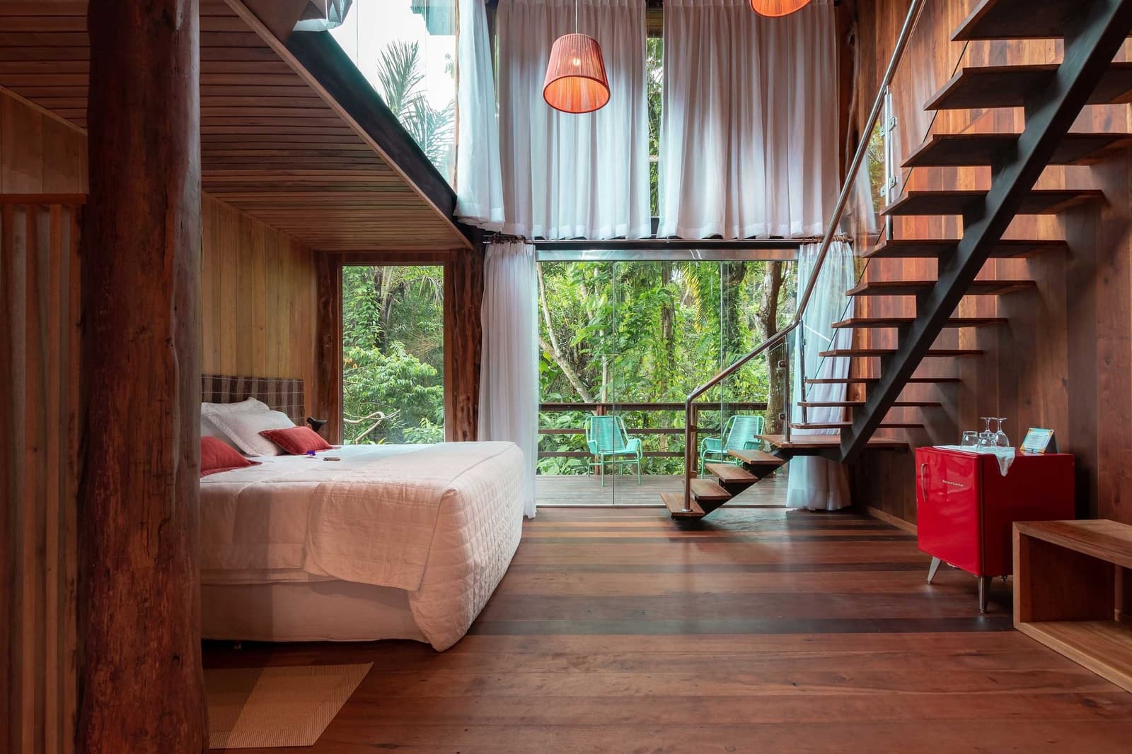 Your bed, the treetops, and nothing else on the agenda. A Mirante do Gavião amazon lodge loft-style suite featuring warm timber walls, a double bed with red accent pillows, a floating steel staircase, sheer white curtains, and floor-to-ceiling glass opening onto a private balcony surrounded by dense rainforest canopy
