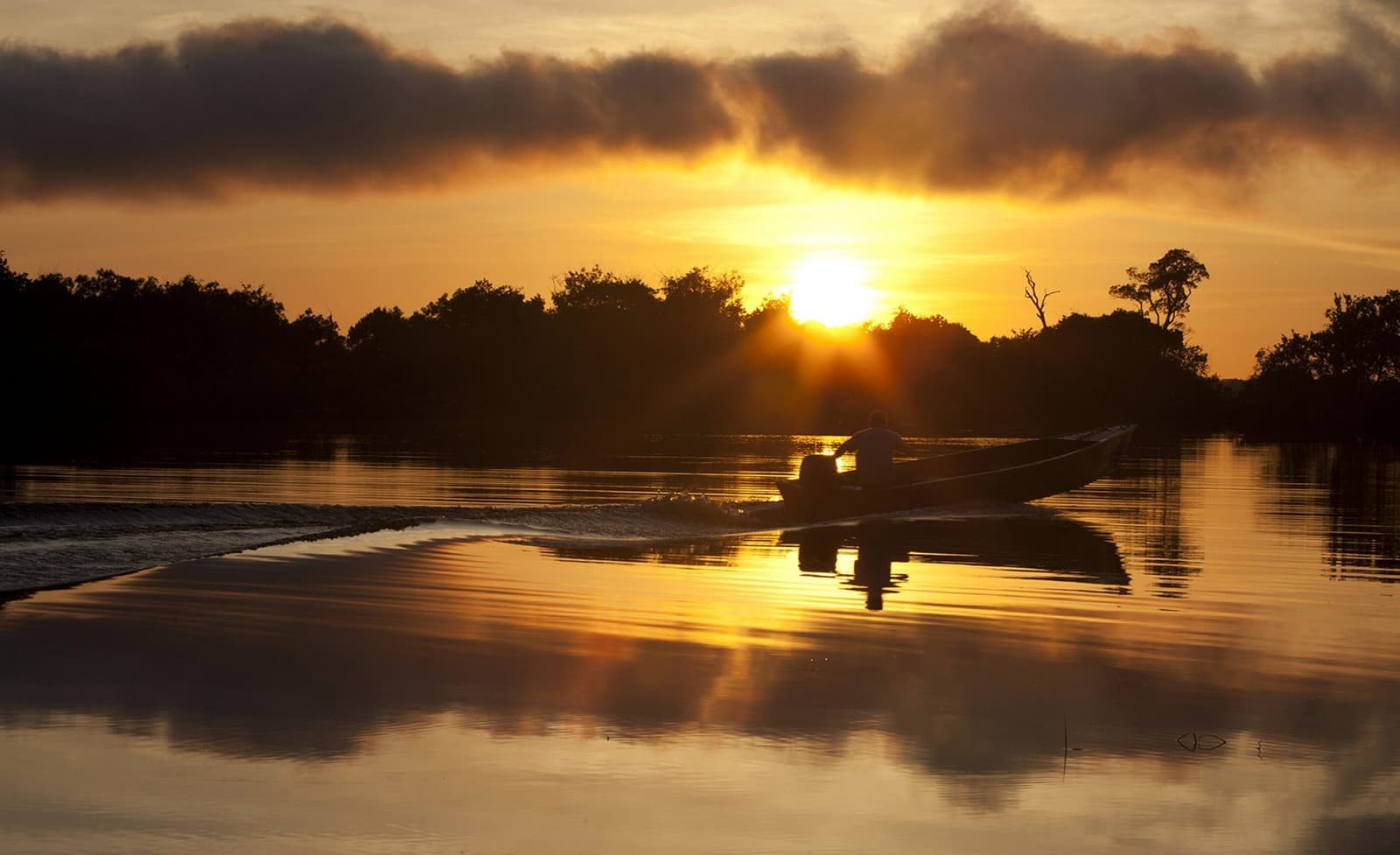 The river keeps its own schedule, and this is the best part of it. A silhouetted figure pilots a motorboat across a glassy river at golden sunset near Mirante do Gavião, with the sun breaking through clouds above a dark treeline