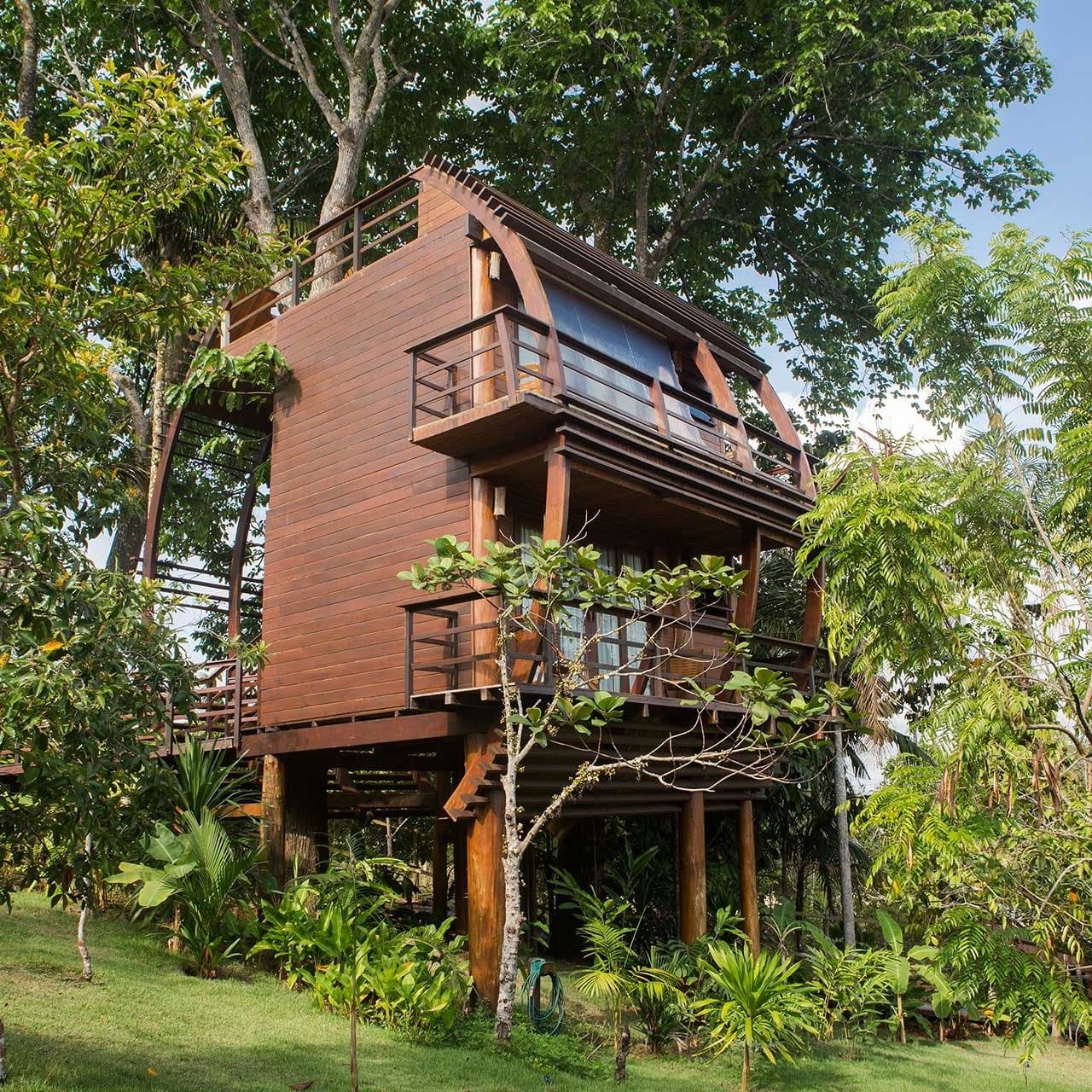 Your room is up there, and yes, the canopy view is even better in person. A multi-storey timber treehouse at Mirante do Gavião rises on stilts above lush tropical gardens, with wraparound balconies on each level and a curved arched roof framed by tall rainforest trees