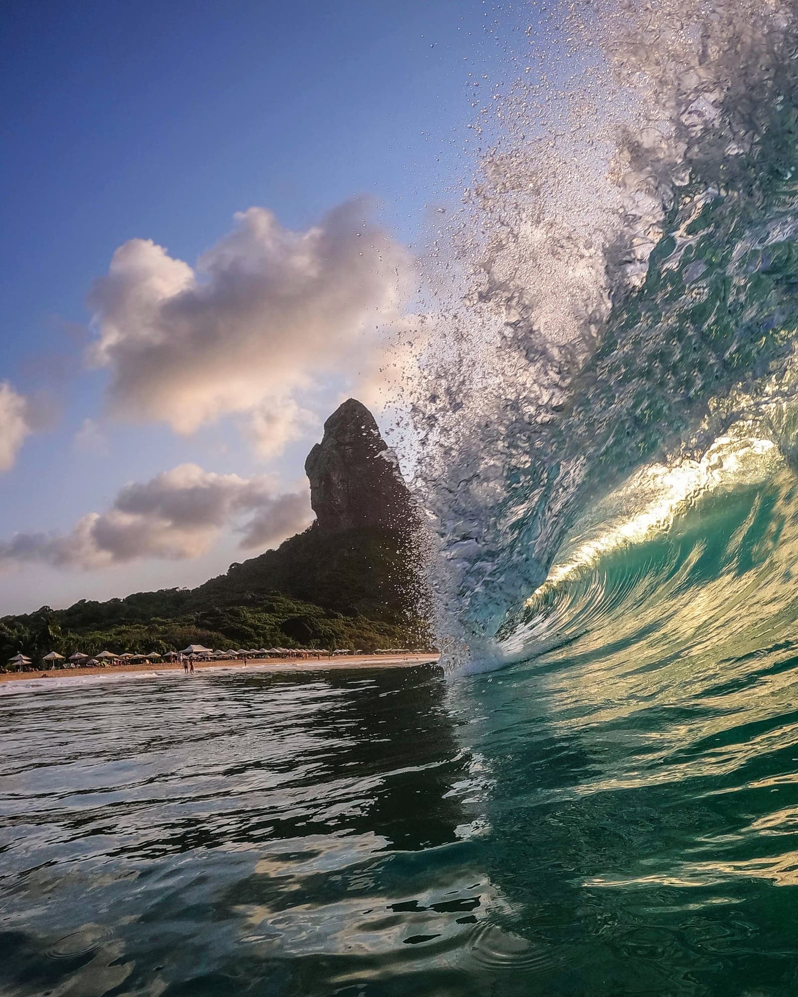 The ocean here has opinions, and it shares them loudly. A turquoise barrel wave curls and splashes in the foreground of an Open Water diving course in Fernando de Noronha, with the iconic Morro do Pico rock pinnacle and a palm-fringed beach visible behind it under a partly cloudy blue sky.