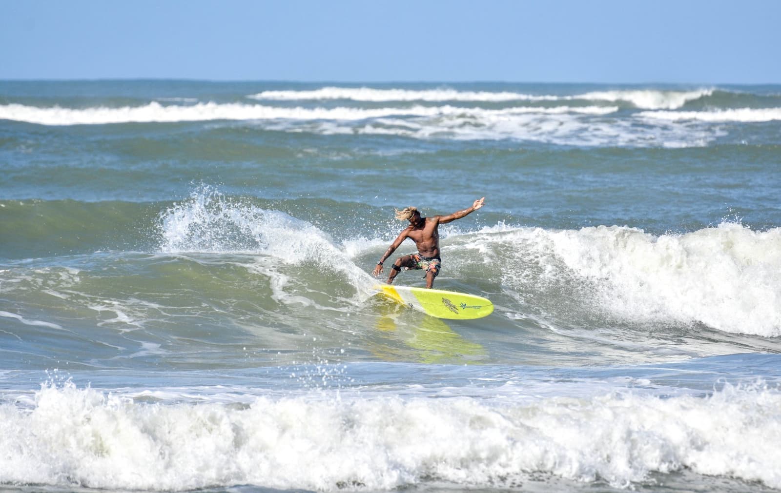 The locals here didn't need a lesson, but watching them is one anyway. Photo: Picasa A local surfer rides a wave on a yellow longboard at a surf school in Itacaré, arms spread wide for balance as white water breaks around him under a clear blue sky.