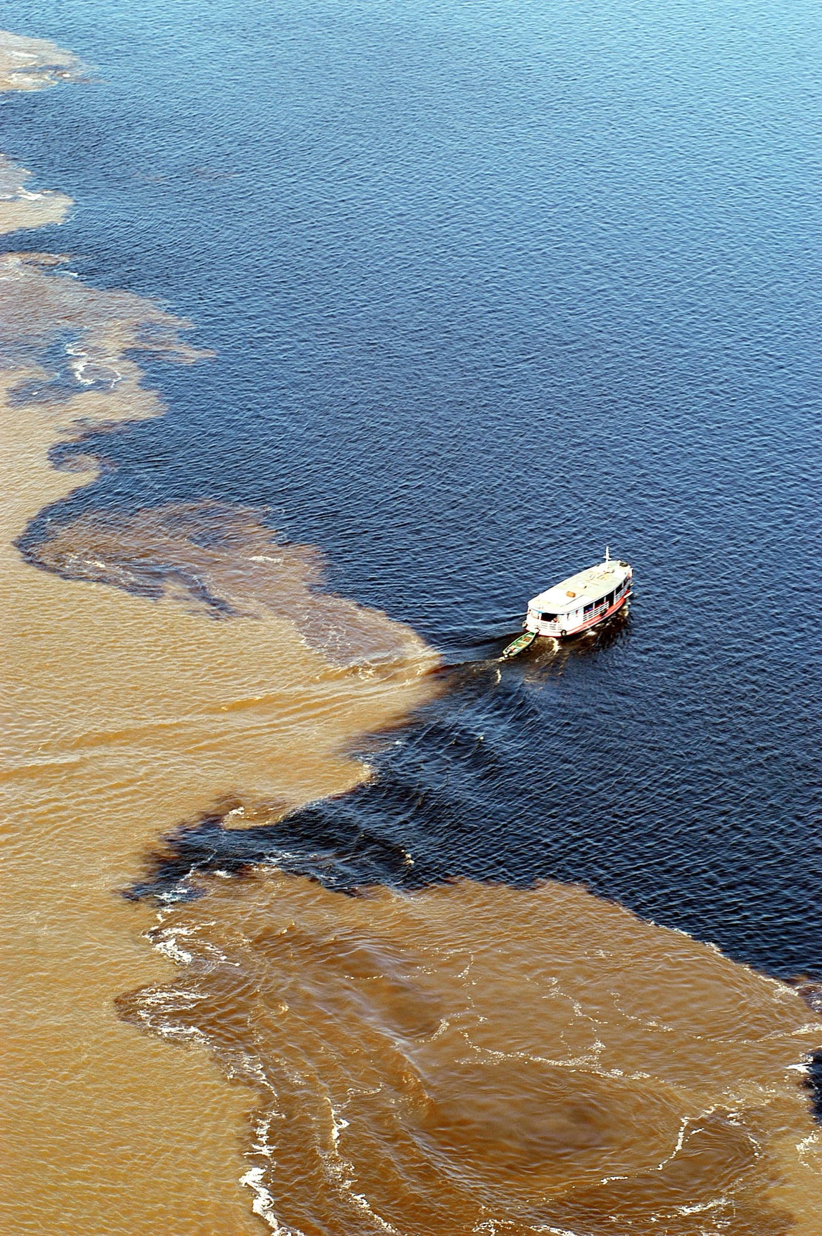 Two rivers, one frame, and they still refuse to share. Aerial view of a river cruise vessel crossing the Meeting of Waters on the tucano amazon river cruise, where dark blackwater and sandy-brown sediment-laden water flow side by side without mixing