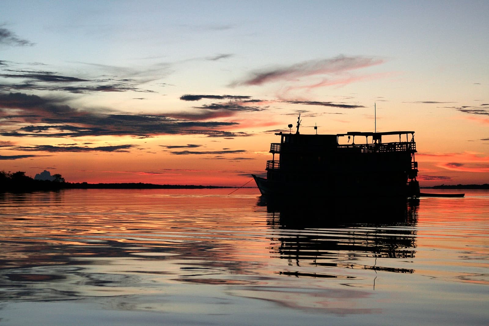 Last light on the river, and the Tucano earns its keep. A multi-deck river vessel silhouetted against a vivid orange and red sunset sky reflected on calm dark water, seen during the Tucano Amazon river cruise