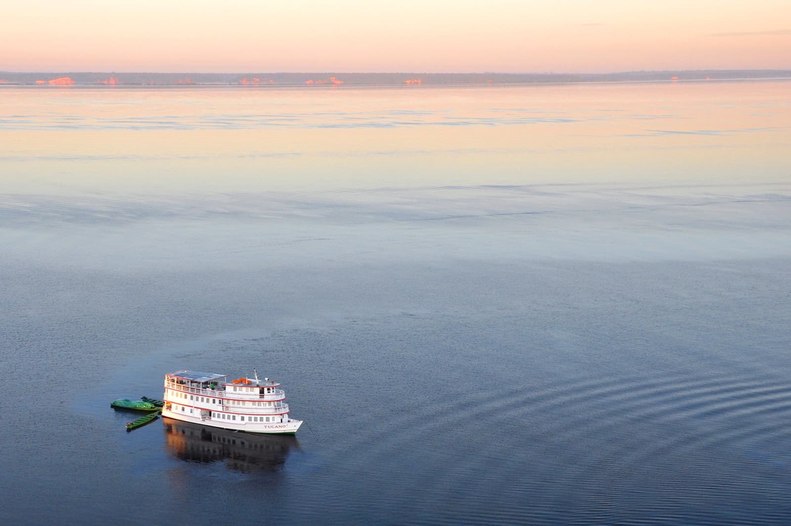 All that water, all yours, and the ship is just getting started. The M/Y Tucano, a small white multi-deck vessel on the Tucano Amazon river cruise, glides across a vast, glassy river at dusk, its wake fanning out behind it beneath a soft pink and peach sky