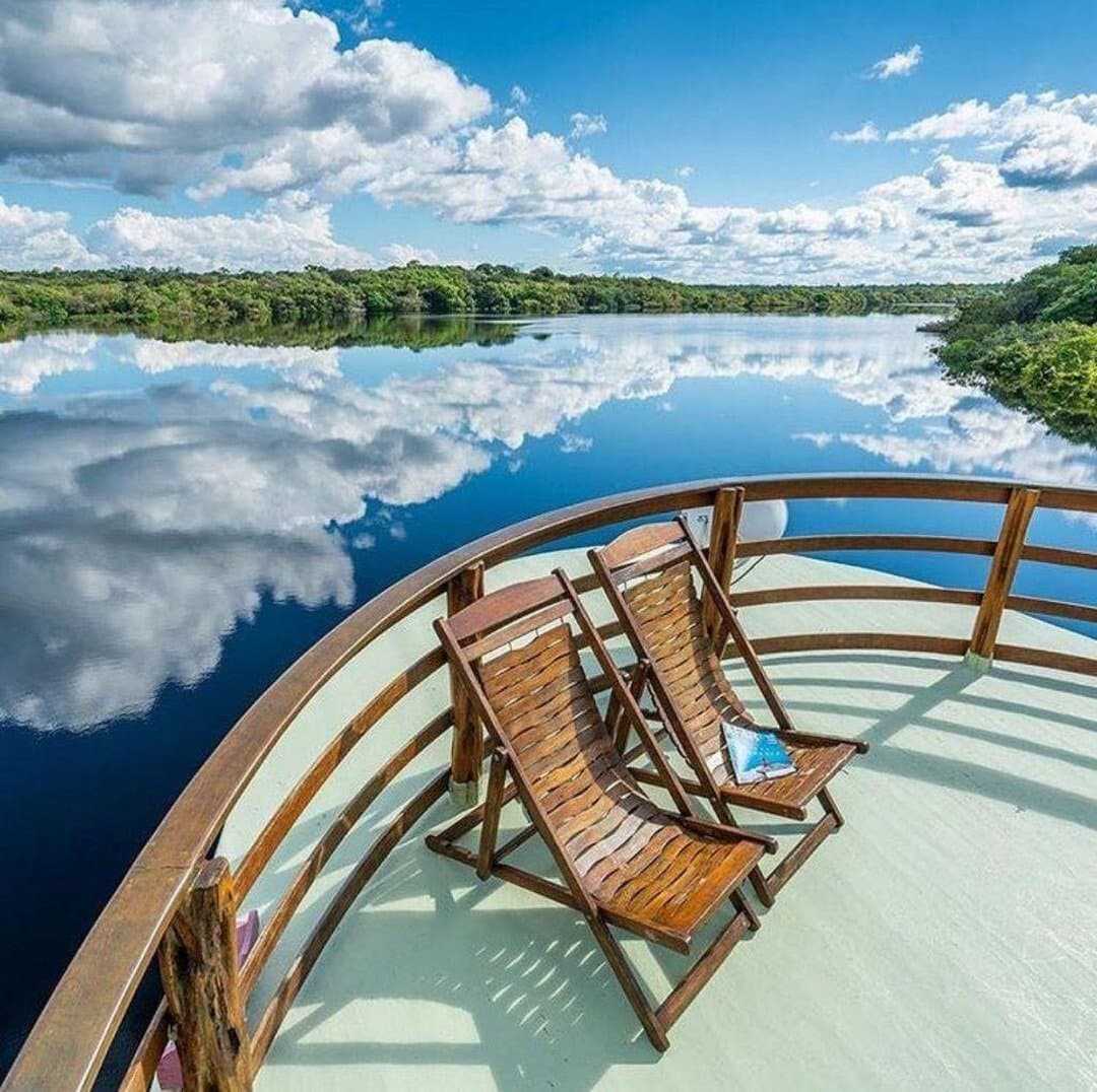 Two chairs, one river, and a sky that can't decide which way is up. Two wooden deck chairs on the curved bow of a riverboat during an Amazon river cruise in Brazil, with clouds perfectly mirrored on a glassy dark-water river flanked by dense rainforest