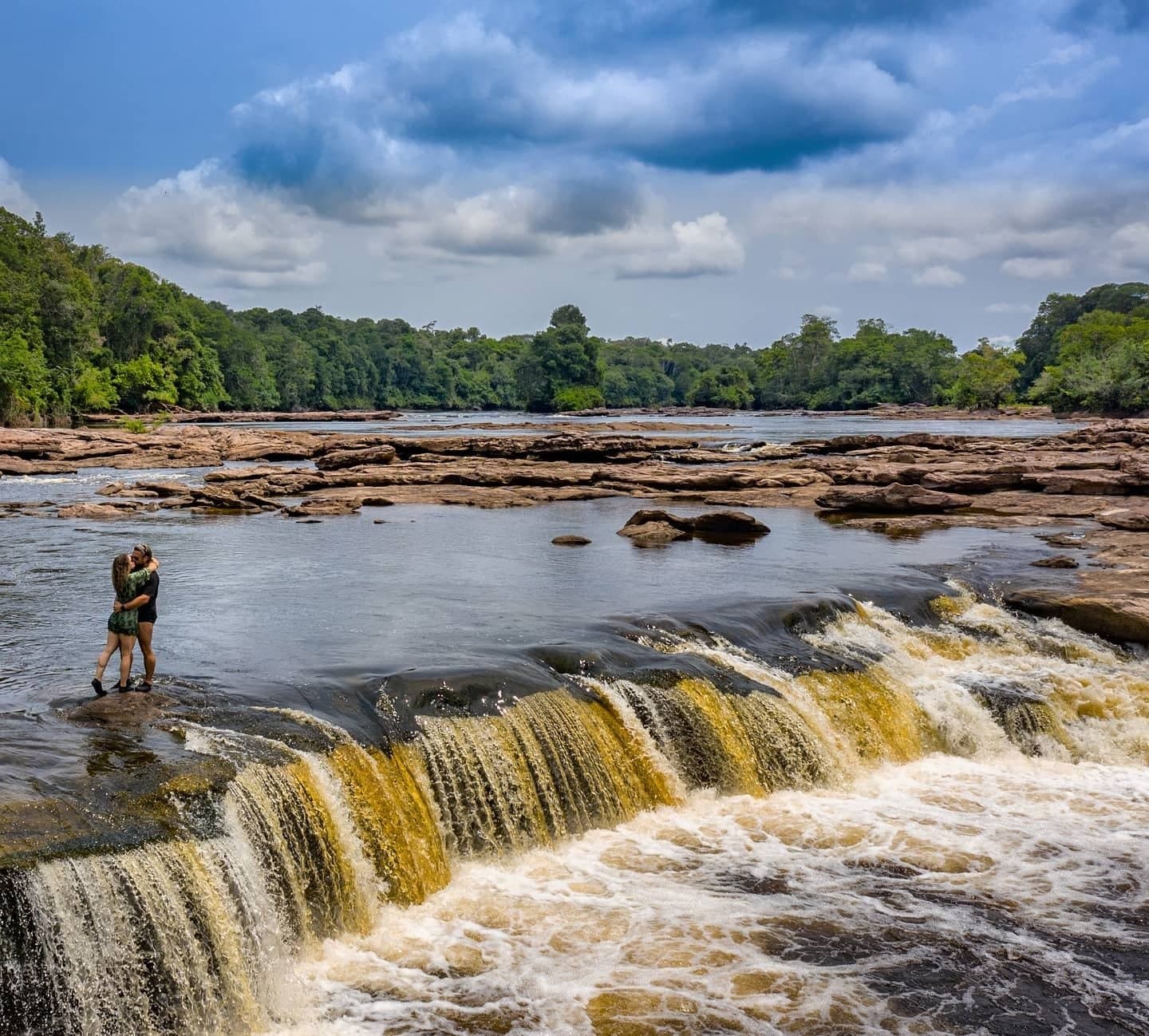 A waterfall this good deserves company. Bring someone worth the trip. A couple embracing on a rock ledge beside a wide, amber-tinted waterfall on an amazon river cruise in Brazil, with flat red sandstone outcrops and dense green forest stretching to the horizon under a dramatic cloudy sky