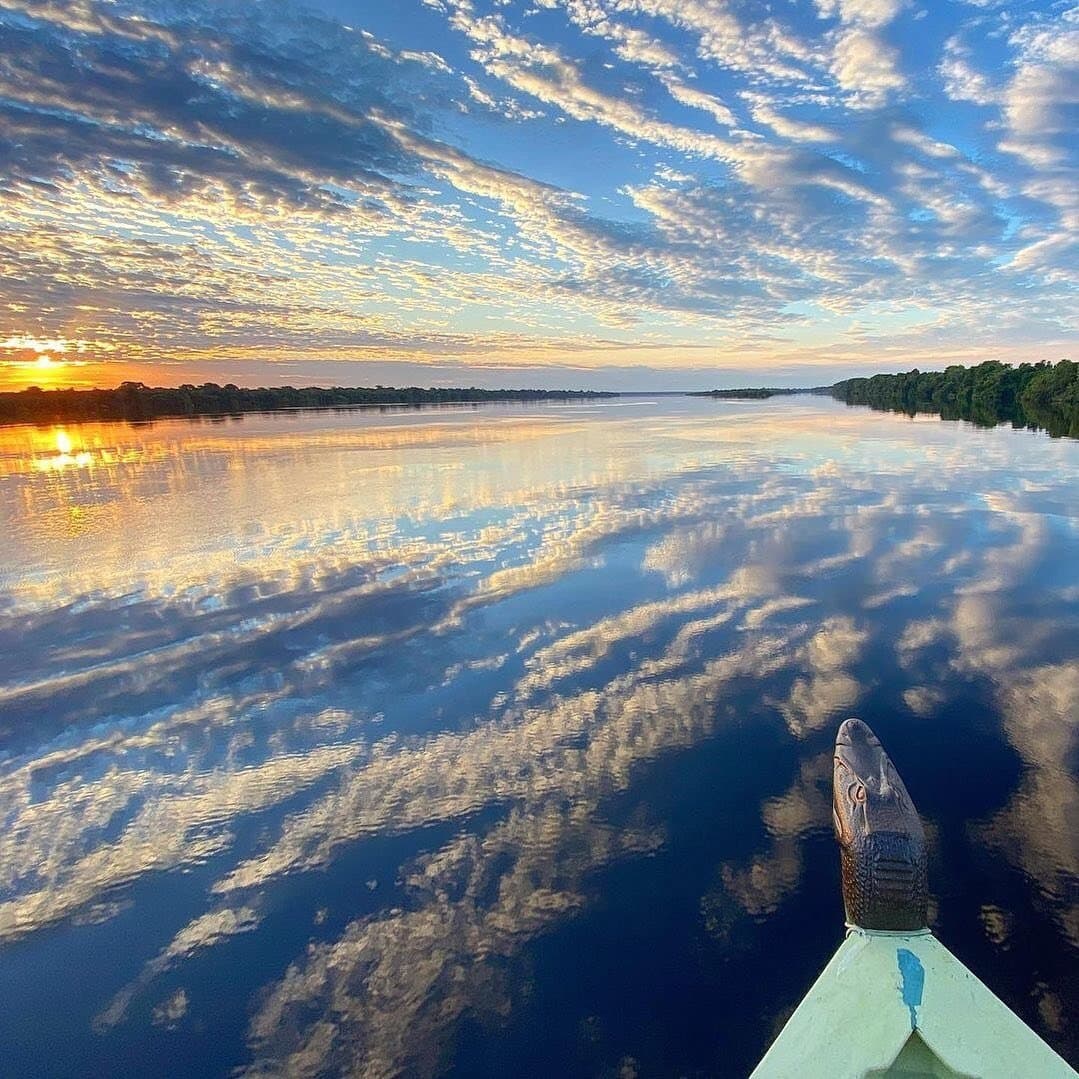 The river doubles the sky, and the boat's figurehead picks the direction. View from the bow of a small riverboat on an amazon river cruise in Brazil, with a carved caiman figurehead pointing toward a glassy black-water river reflecting orange sunrise clouds and flanked by dense forest on both banks