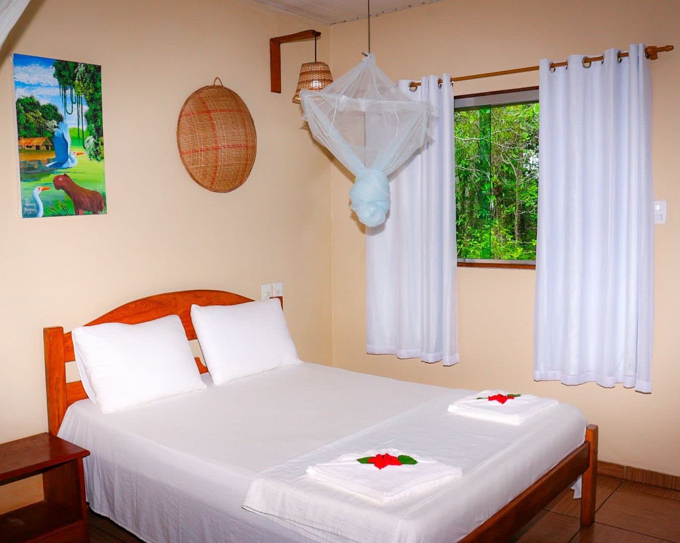 Folded towels, fresh flowers, and the forest just outside the window, sleep well tonight. A cozy Amazon Turtle Lodge bedroom with a wooden-frame double bed, white mosquito net canopy, red hibiscus flower towel decorations, wicker wall art, and a window overlooking dense green rainforest