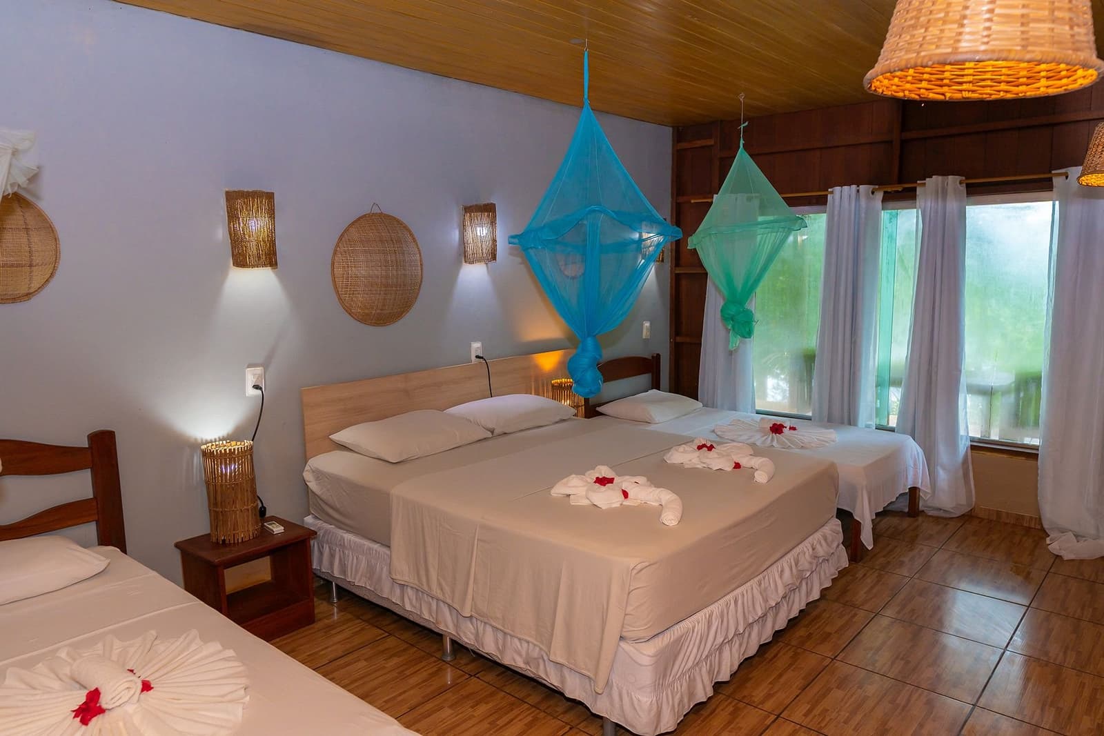 Towel swans, canopy nets, and the jungle just outside the window. Sleep sorted. A triple bedroom at the Amazon Turtle Lodge with colourful hanging mosquito nets, folded towel decorations on crisp white linens, woven rattan wall lamps, and a large window overlooking lush greenery