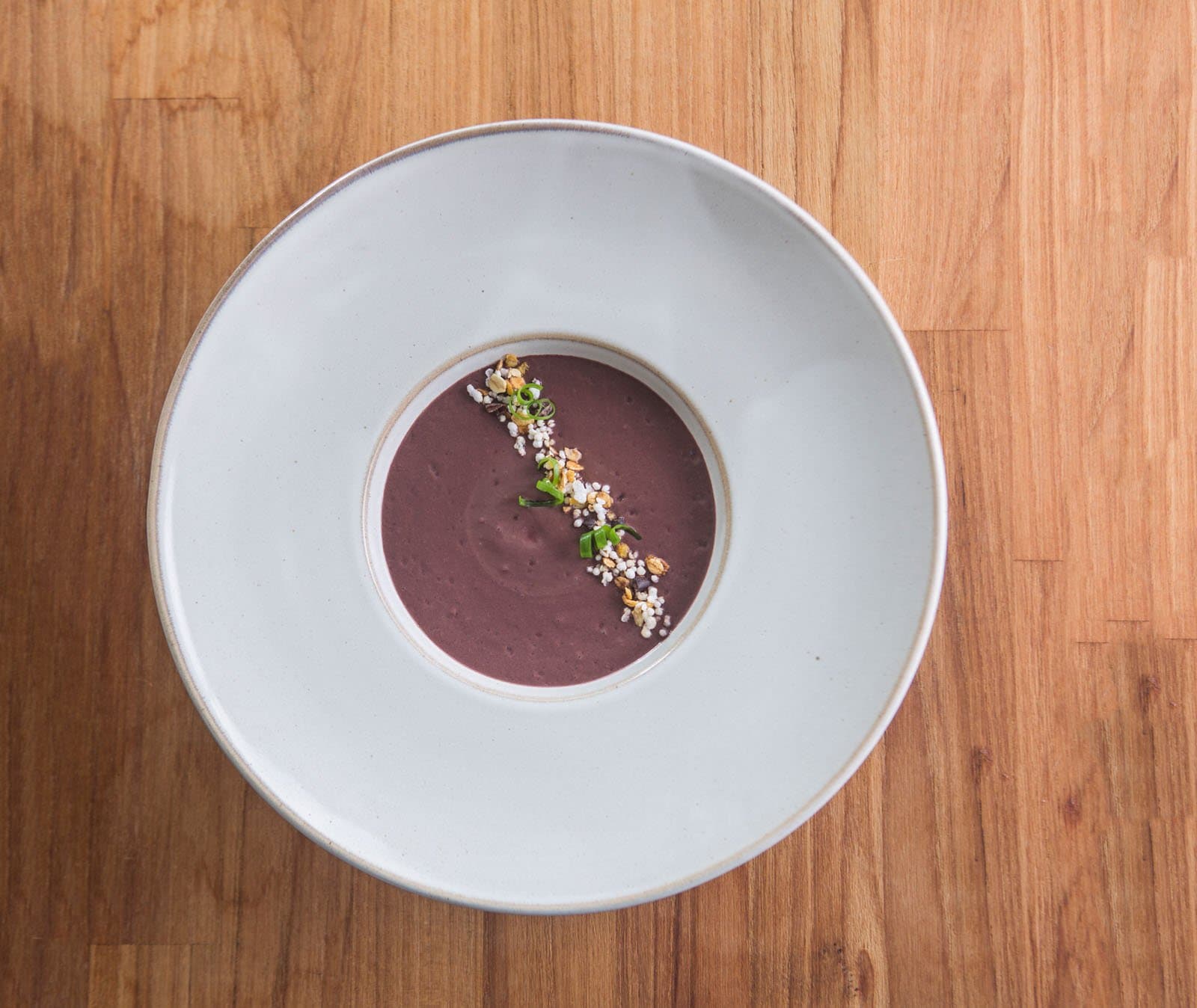 Simple ingredients, serious kitchen. The forest earns you this. Photo: Rjcastilho Overhead view of a smooth deep-purple cream soup served in a wide white ceramic bowl, garnished with a diagonal line of toasted grains, puffed rice, and sliced green onion, on a warm wood surface at a birdwatching in the amazon rainforest lodge