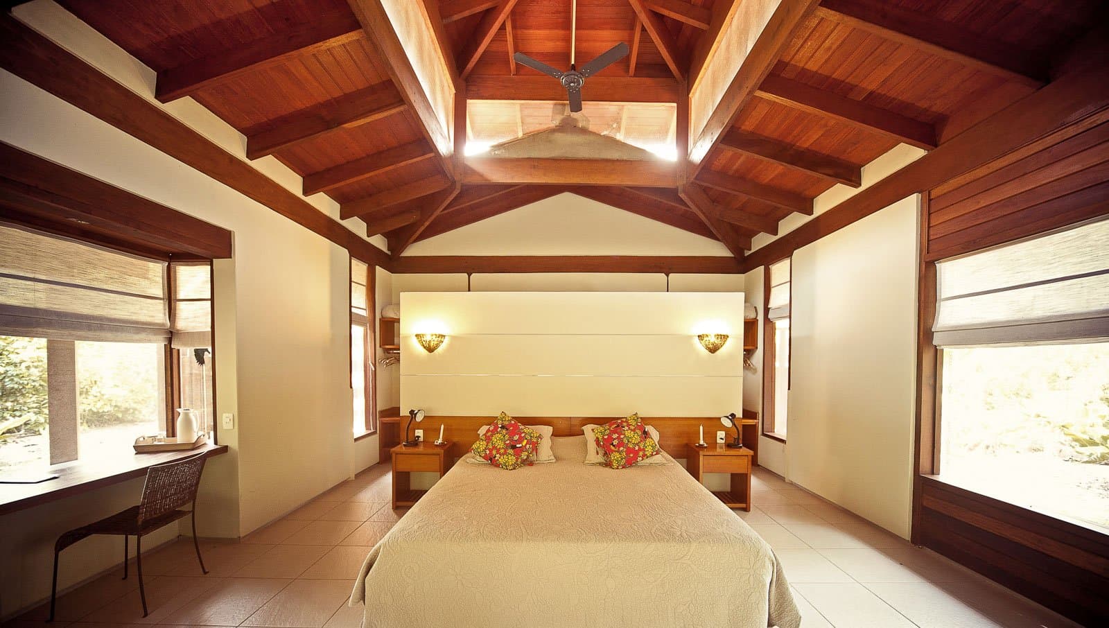 Sleep well tonight. The forest starts early and it won't wait. Photo: Samuel Melim Spacious lodge bedroom with a vaulted timber ceiling, ceiling fan, double bed with floral cushions, warm wall sconces, and windows overlooking green forest — ideal base for birdwatching in the Amazon rainforest