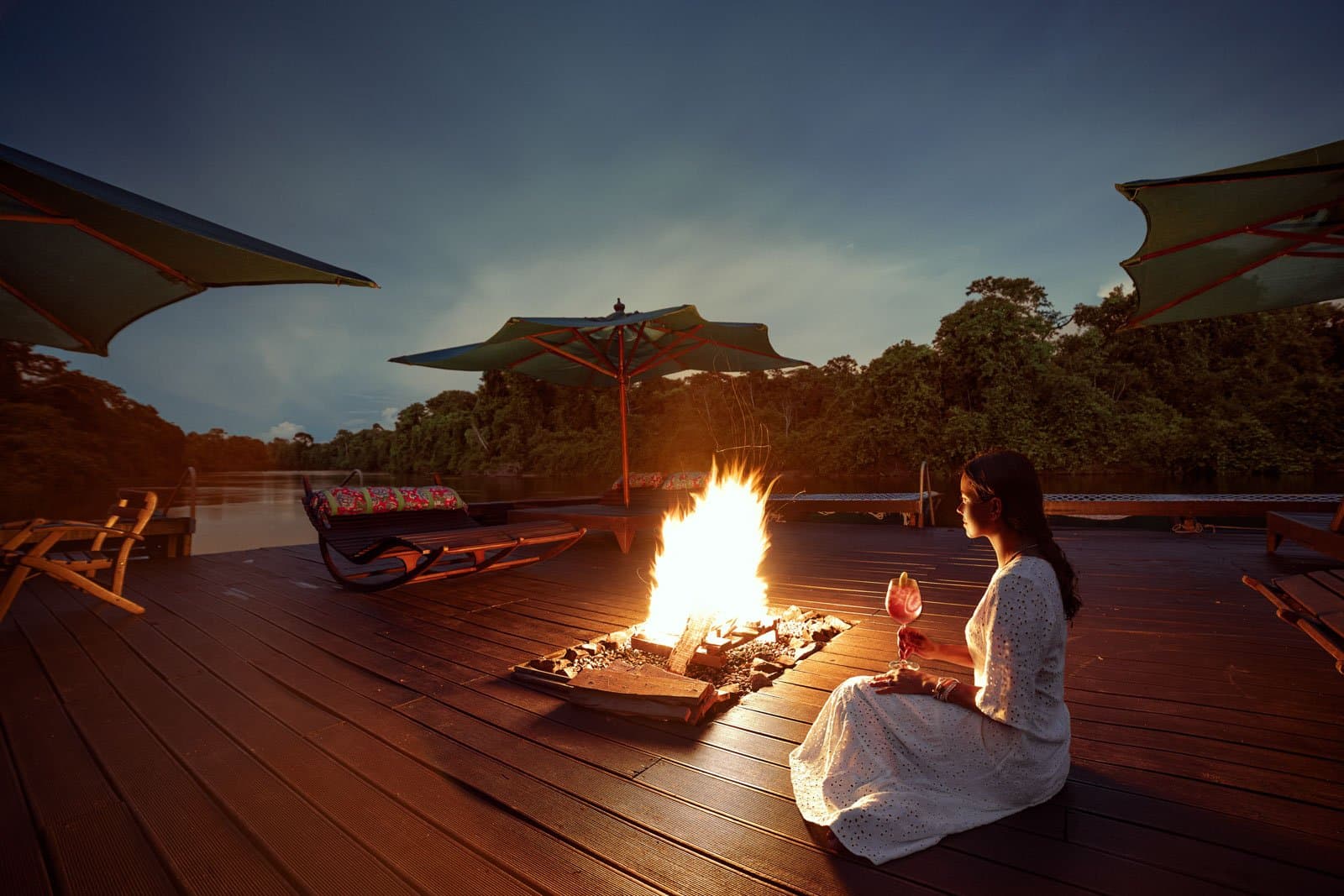 Cocktail in hand, bonfire lit, and the forest settling in for the night. Photo: Samuel Melim A woman in a white dress sits beside an open fire pit on a wooden deck overlooking a jungle river at dusk, holding a cocktail at a birdwatching in the amazon rainforest lodge