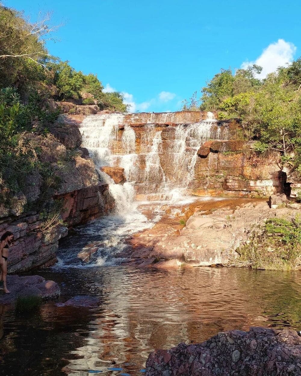 Cold water, warm rock, no agenda. This is the Chapada at its most honest. A tiered waterfall cascading over rust-colored sandstone ledges into a clear natural pool on the Chapada Diamantina Tour, with a visitor wading at the water's edge and green cerrado vegetation under a blue sky