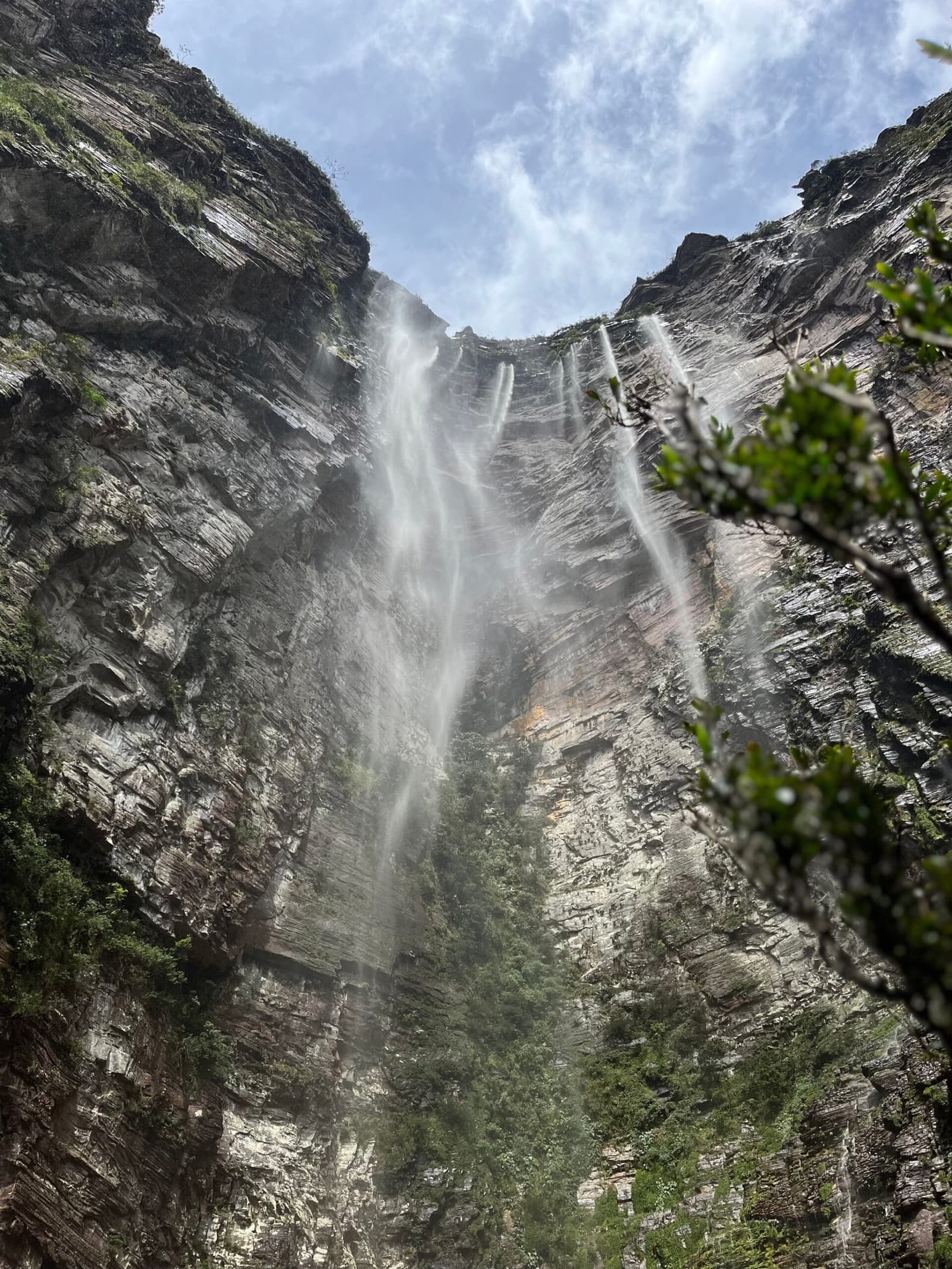 All that height, and the mist still finds you at the bottom. Photo: Lucas Neves Upward view from the base of Cachoeirão waterfall on a Chapada Diamantina trek, with multiple streams of water trailing down a towering layered sandstone cliff face framed by green vegetation and a partly cloudy sky