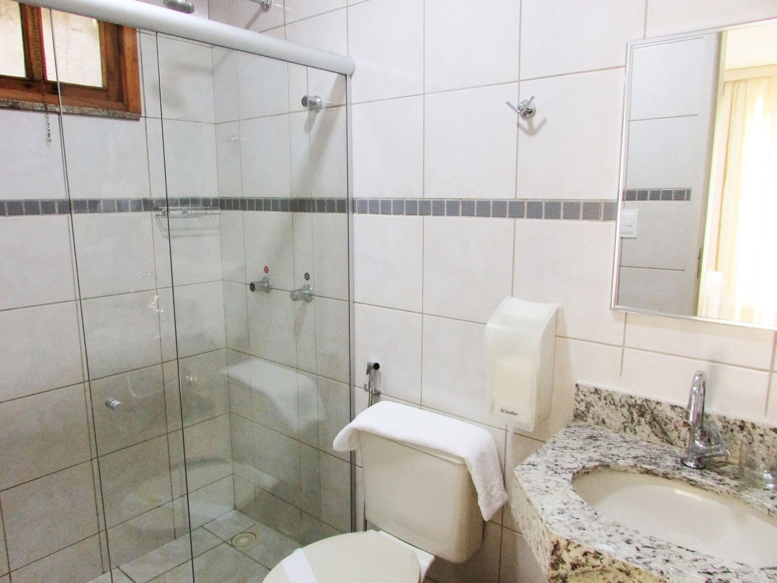 After 30 km in the saddle, a hot shower feels earned. Clean private en-suite bathroom in a cycling Bahia guesthouse, featuring a glass-enclosed shower, granite vanity, and white tiled walls with a decorative border strip