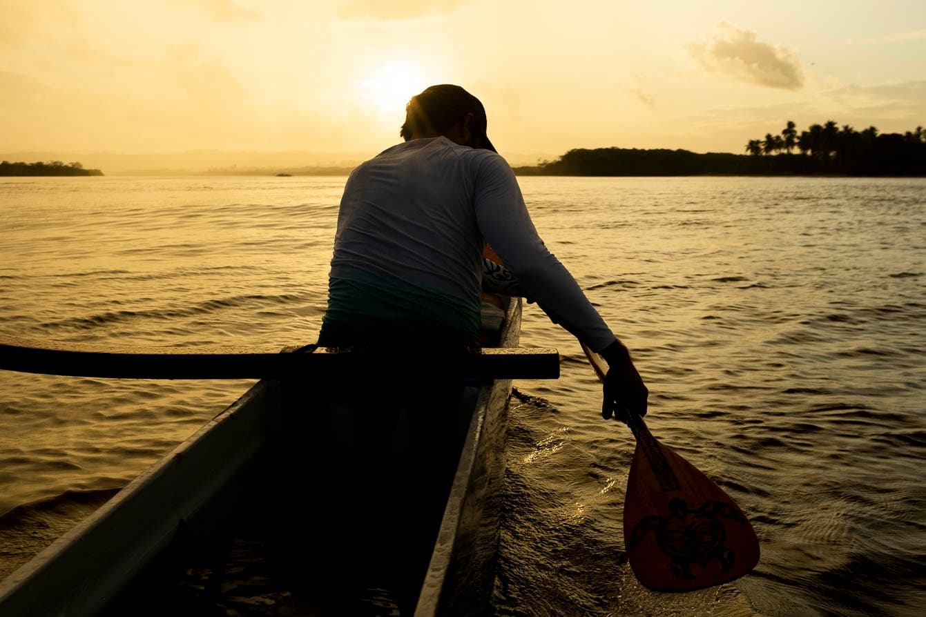 Paddle in hand, the river does the rest. A paddler in a Hawaiian canoe dips his wooden oar into calm water on a cycling bahia tour, silhouetted against a golden sunset sky with a forested shoreline in the distance