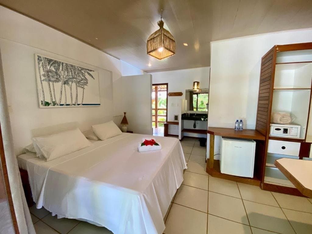 Clean, quiet, and yours for the night. Tomorrow the coast calls. Bright double room at an Itacaré guesthouse on a cycling Bahia tour, with white bedding, a woven pendant lamp, palm-tree wall art, mini fridge, and an open door leading to a garden terrace