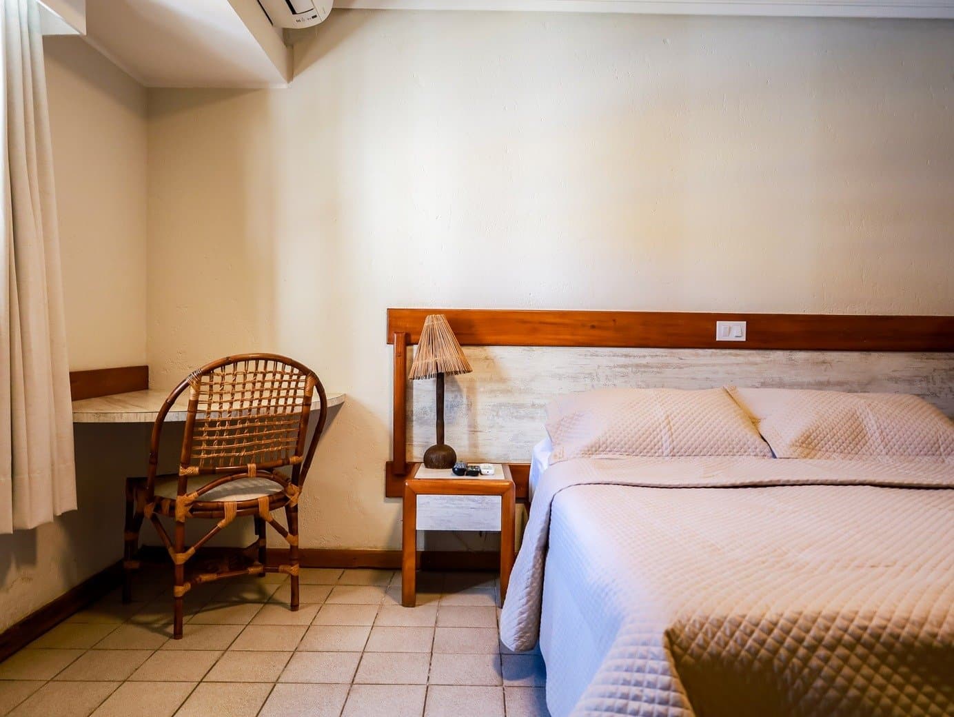 Unpretentious, clean, and exactly what tired legs ask for. Simple, comfortable guesthouse room on a cycling Bahia tour, featuring a double bed with light quilted covers, a rattan chair, wooden headboard, and a woven bamboo table lamp