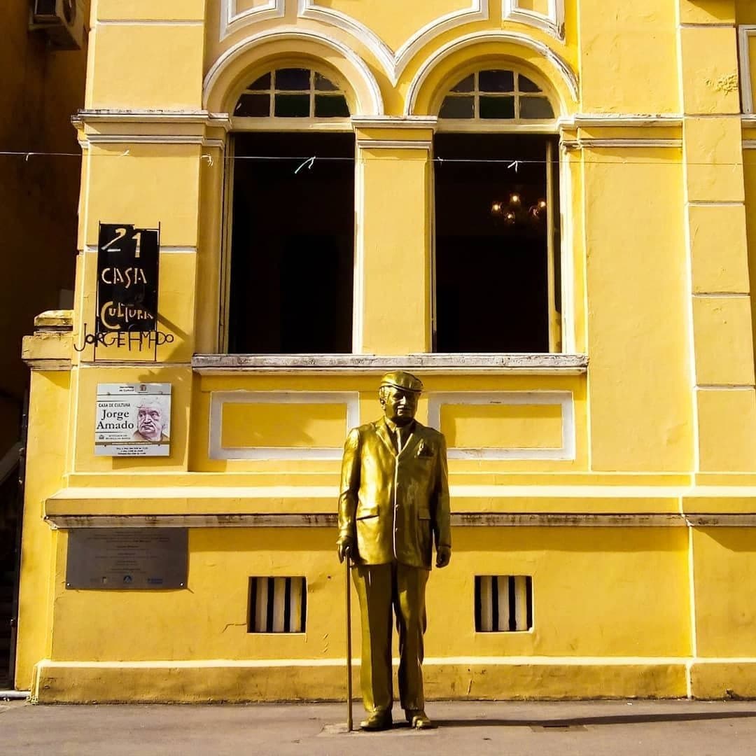 He wrote the whole region into legend. This is where the story starts. Gold statue of Jorge Amado with a walking cane standing before the bright yellow facade of the Casa de Cultura Jorge Amado in Ilhéus, Bahia Brazil