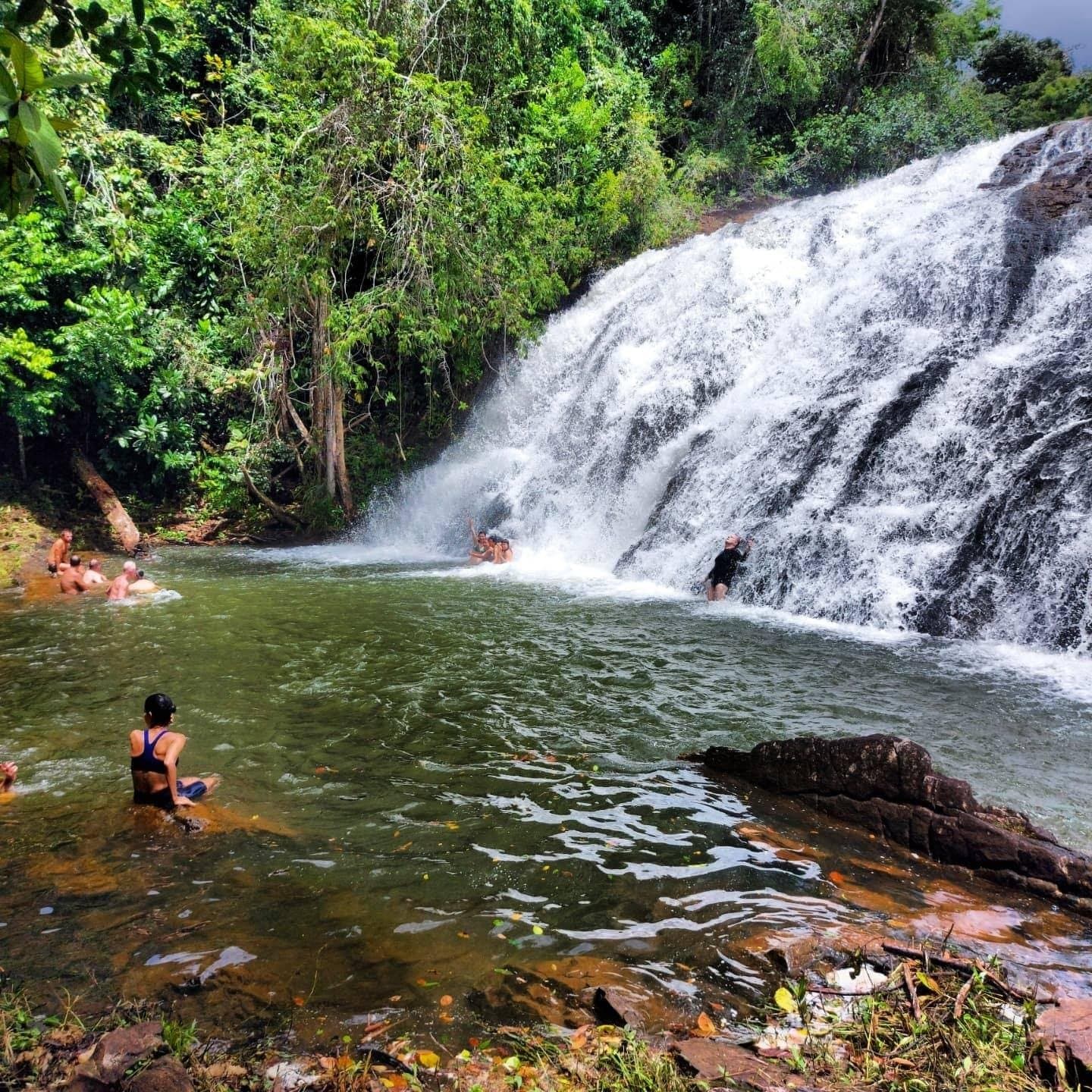 Bikes parked, shoes off, everyone in. Some rewards you have to swim to. Travelers swimming in the natural pool at Milagrosa Waterfall, a wide cascade tumbling over dark rock into a green pool surrounded by Atlantic Forest, a reward on the cycling Bahia cocoa route