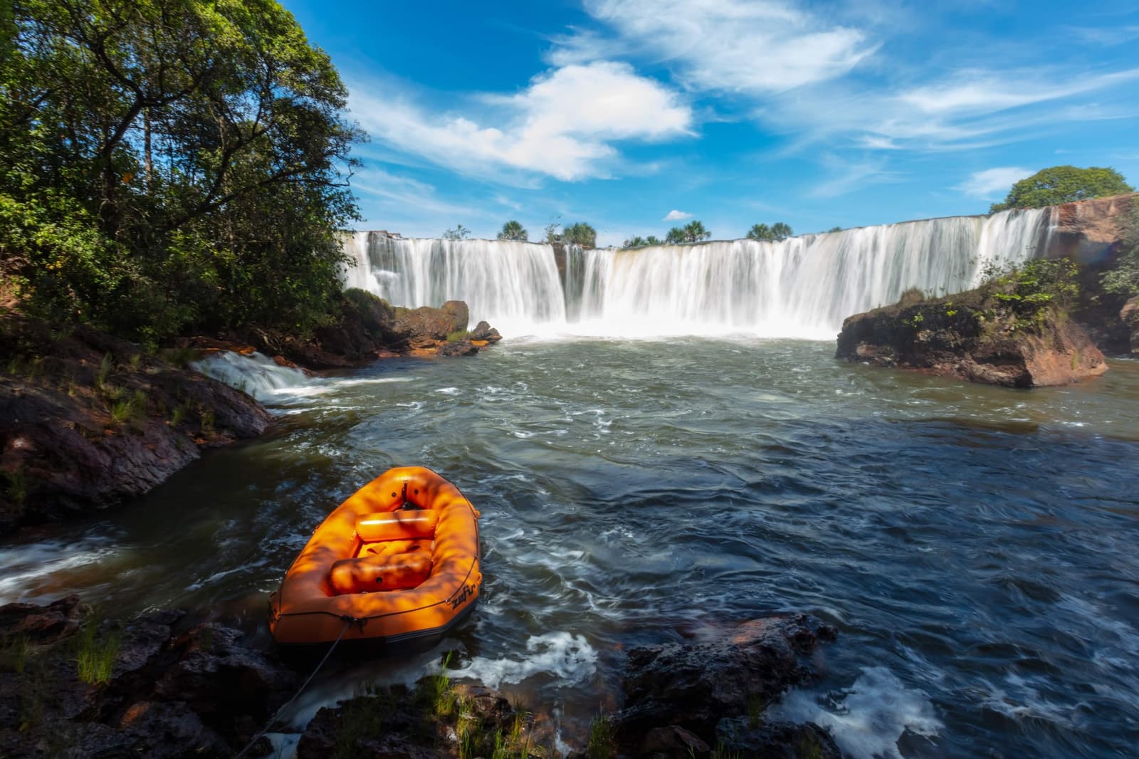 Lunch first, then you float straight toward that curtain of water. Photo: Alberto Andrich An orange inflatable raft moored on rocky riverbanks in front of a wide horseshoe-shaped waterfall during a Jalapão trekking expedition, surrounded by cerrado forest under a blue sky