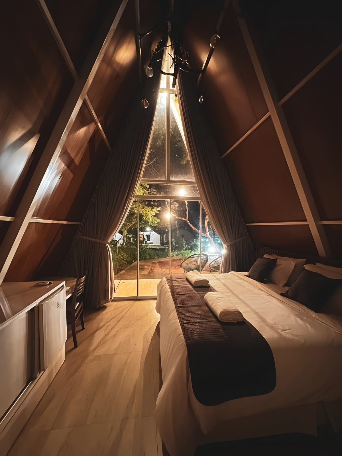 Good beds matter more after a day of hiking through Cerrado. This one delivers. Interior of a wooden A-frame cabin at night during a Jalapão trip, with a king bed dressed in white linens, a tall triangular glass window overlooking an illuminated garden, and warm Edison-bulb lighting overhead
