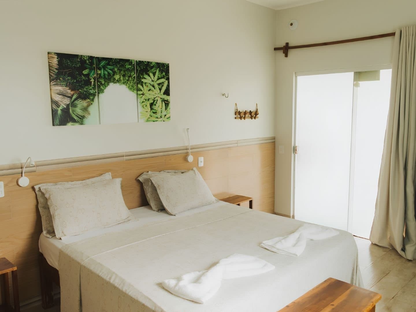 Rest well tonight. The dunes and fervedouros are waiting for you tomorrow. Bright double room at a Jalapão trip guesthouse, featuring a wood-panelled headboard, crisp white bedding with folded towels, tropical nature wall art, and a sliding door opening to natural light