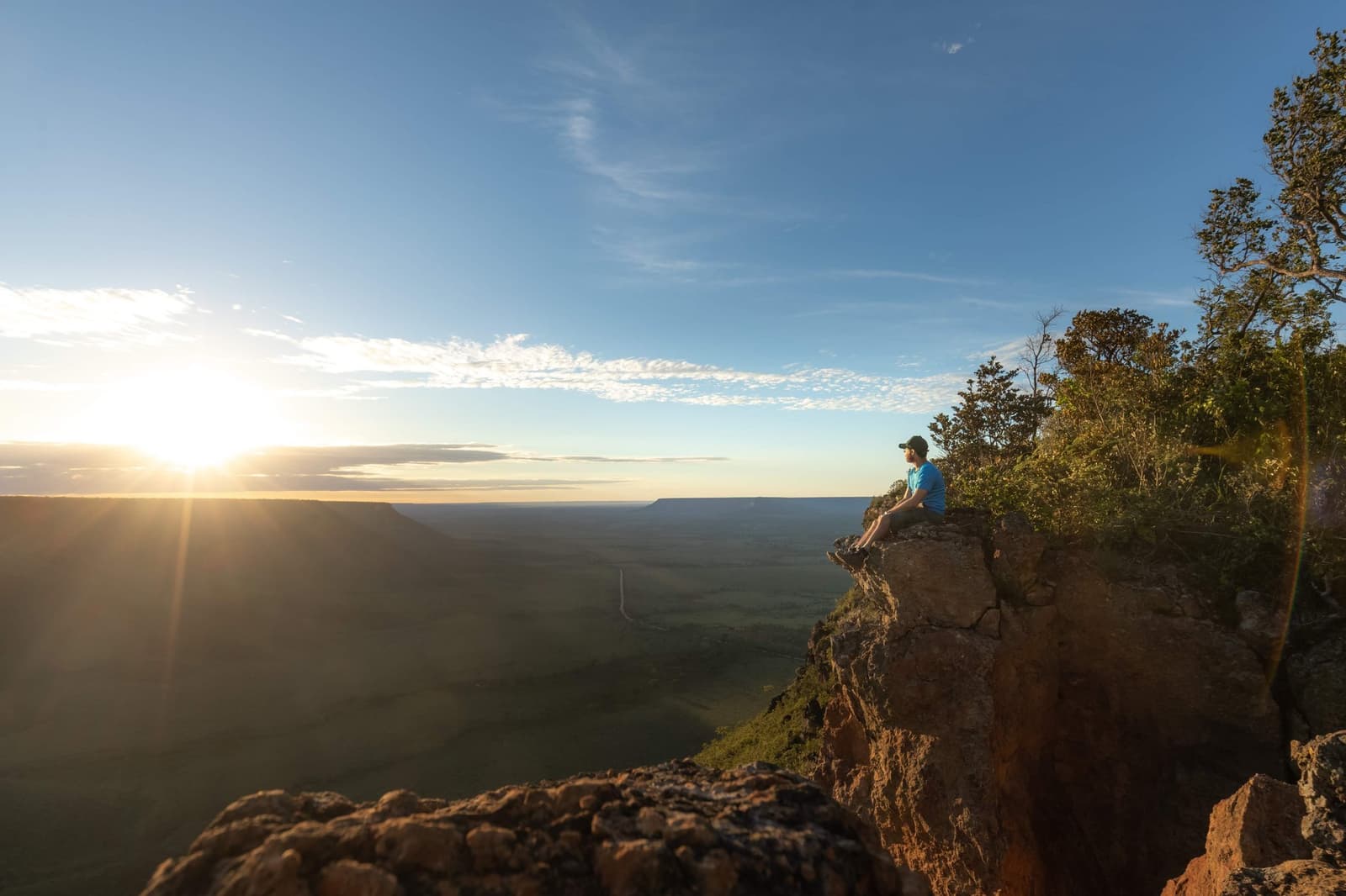 Up before dawn, perched on rock, watching the light arrive. Worth every early alarm. Photo: Alexandre Socci A traveler sits on a sandstone cliff edge at sunrise during a Jalapão trip, looking out over a vast green plateau and distant mesa under a wide blue sky