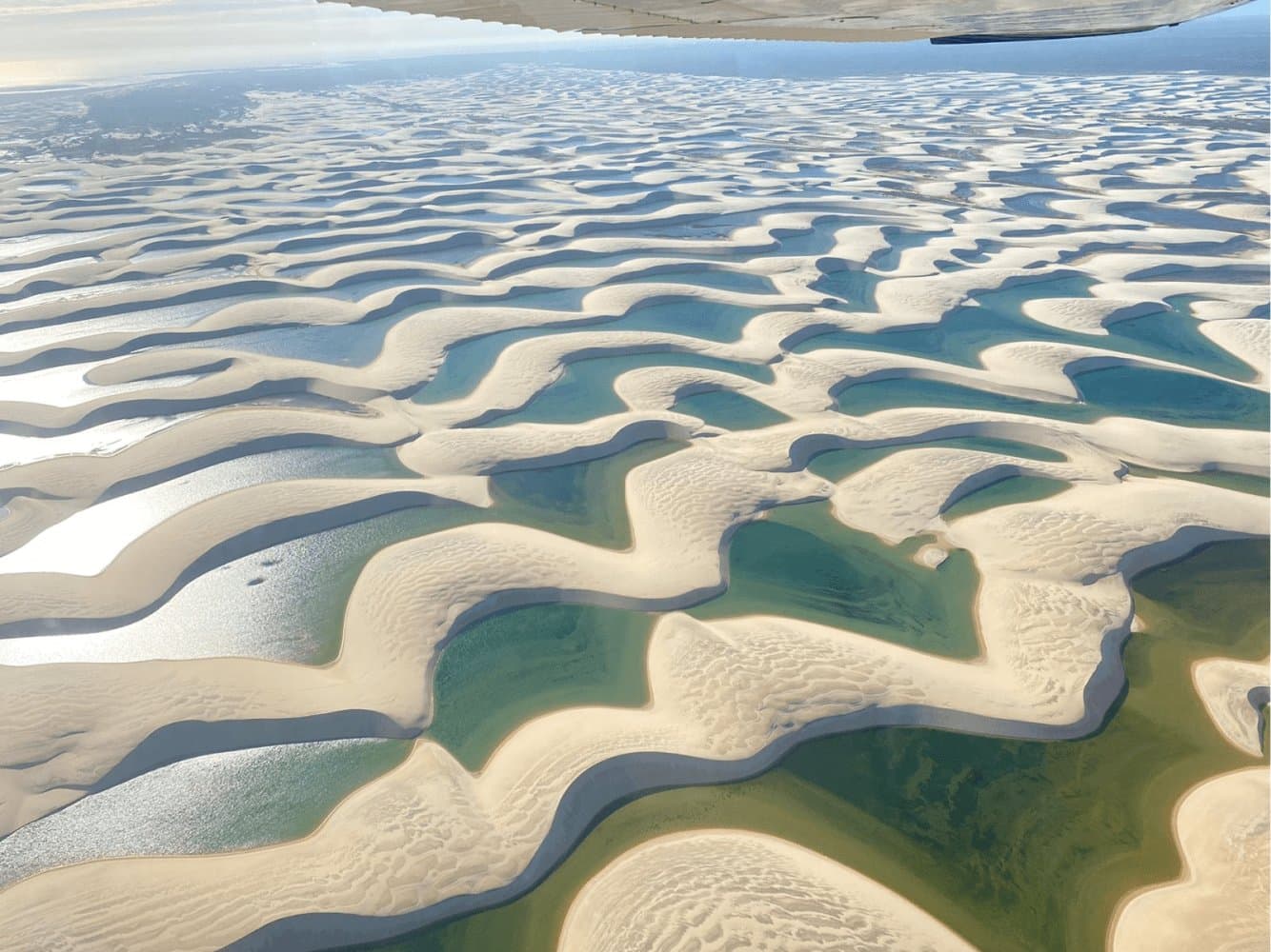 From up here, the dunes and lagoons look like brushstrokes on a canvas nobody planned. Aerial view from a small aircraft over the Lençóis Maranhenses full itinerary landscape, showing vast white sand dunes interlaced with turquoise and emerald seasonal lagoons stretching to the horizon