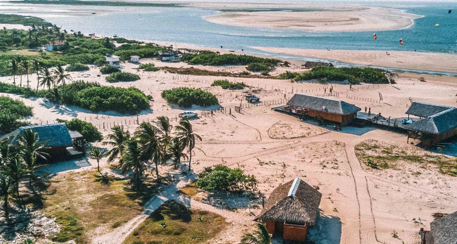 Life here runs on sand, breeze, and very little else. Aerial view of thatched-roof huts, palm trees, and sandy tracks in the rustic village of Atins, with the Lençóis Maranhenses Full Itinerary taking travelers along the coastline where kite surfers dot the blue waters beyond