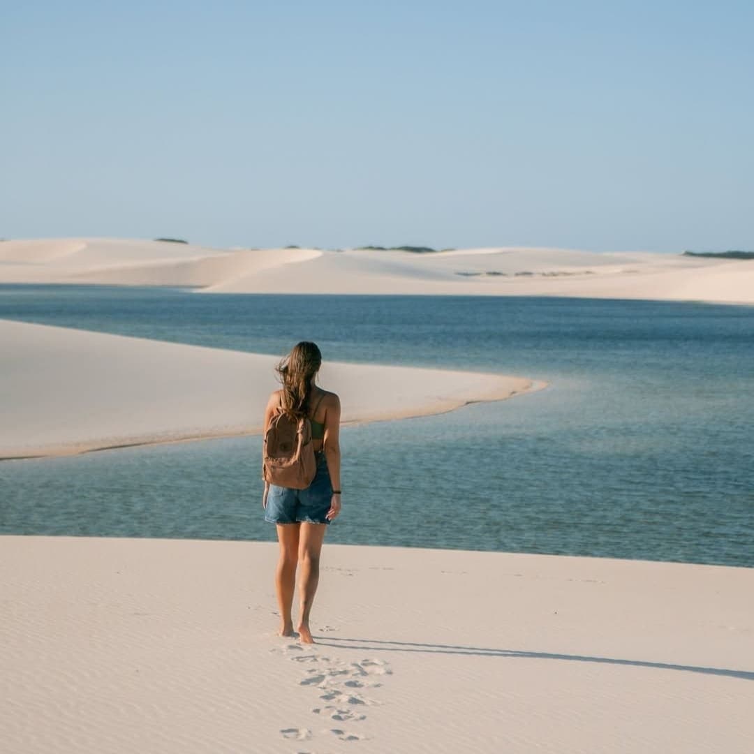 Footprints in the sand, a lagoon ahead, no agenda required. A woman with a backpack walks barefoot across white sand dunes toward a crystal-clear blue lagoon on a Lençóis Maranhenses full itinerary, her footprints trailing behind her