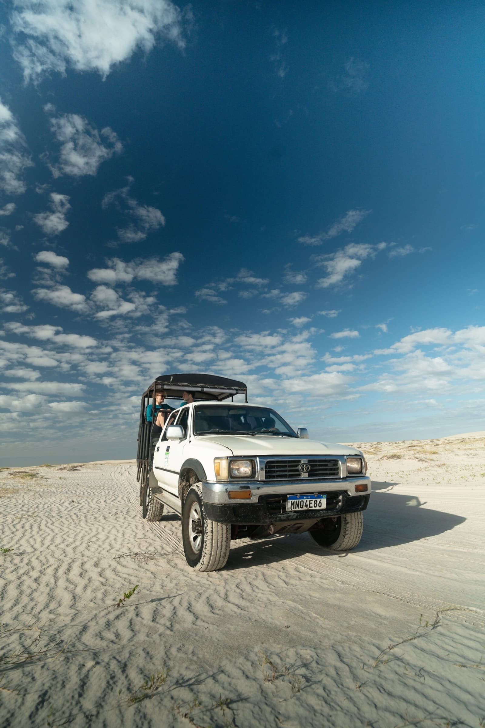 Your ride across one of Brazil's most surreal landscapes, no road required. A white Toyota 4x4 pickup with an open canopy bed carrying passengers crosses a vast expanse of pale white sand dunes under a deep blue sky with scattered clouds on the lencois maranhenses trek.