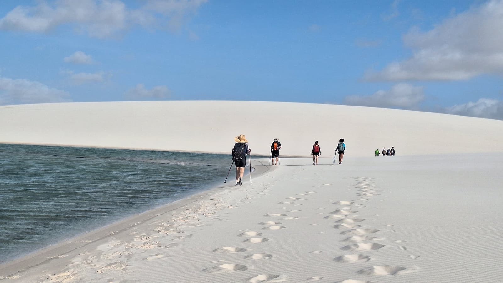 Sand on one side, water on the other, the trail picks itself. A group of trekkers with hiking poles and backpacks walks along a narrow white sand ridge beside a dark-green freshwater lagoon during the Lençóis Maranhenses trek from Atins, with a vast pale dune curving against a blue sky ahead