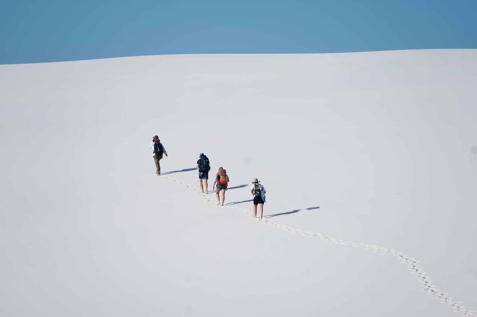Four people, one enormous dune, and a trail that belongs only to them. Four hikers with backpacks walking single-file up a vast white sand dune on the Lençóis Maranhenses trek, leaving a trail of footprints across the empty slope under a clear blue sky